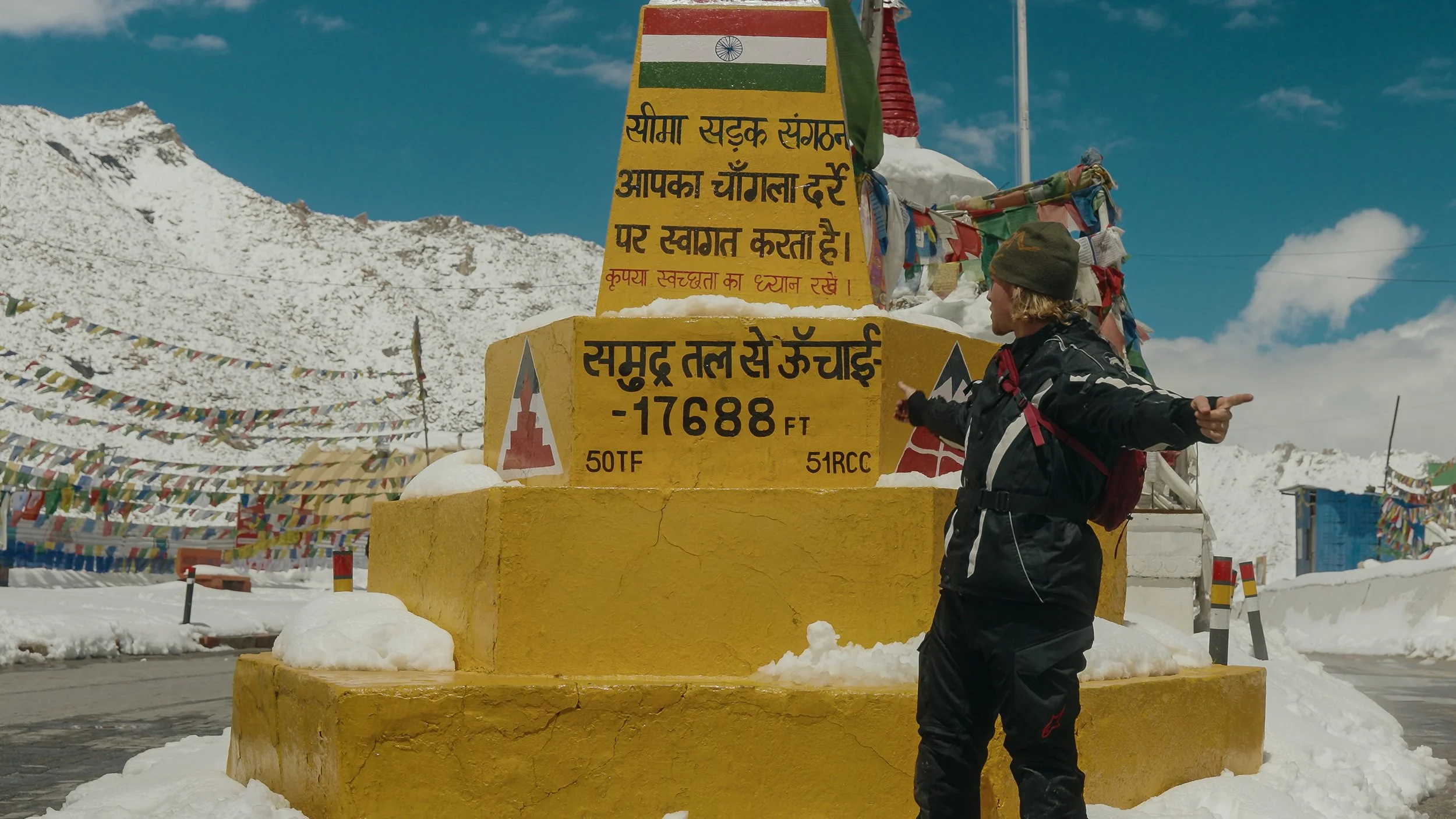 A person wearing a green beanie and black jacket standing in front of a yellow sign with Hindi and English writing, indicating an elevation of 16,888 feet at a mountain pass, with mountain peaks and prayer flags in the background.