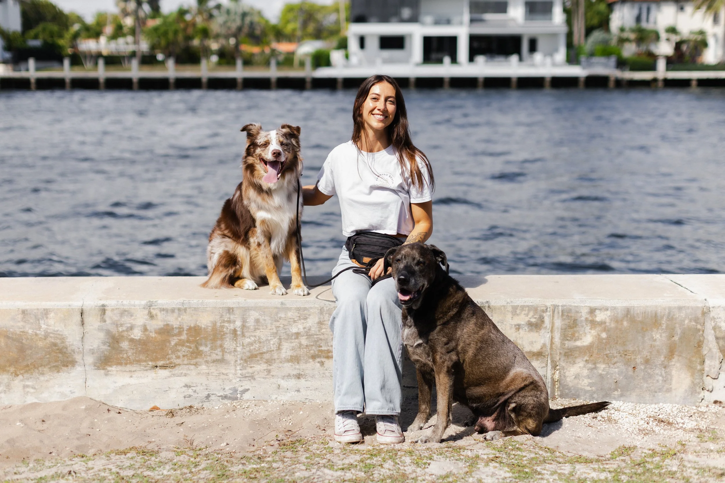 A woman sitting on a concrete ledge near water, smiling, with two dogs, one Australian Shepherd and one mixed breed, enjoying a sunny day.
