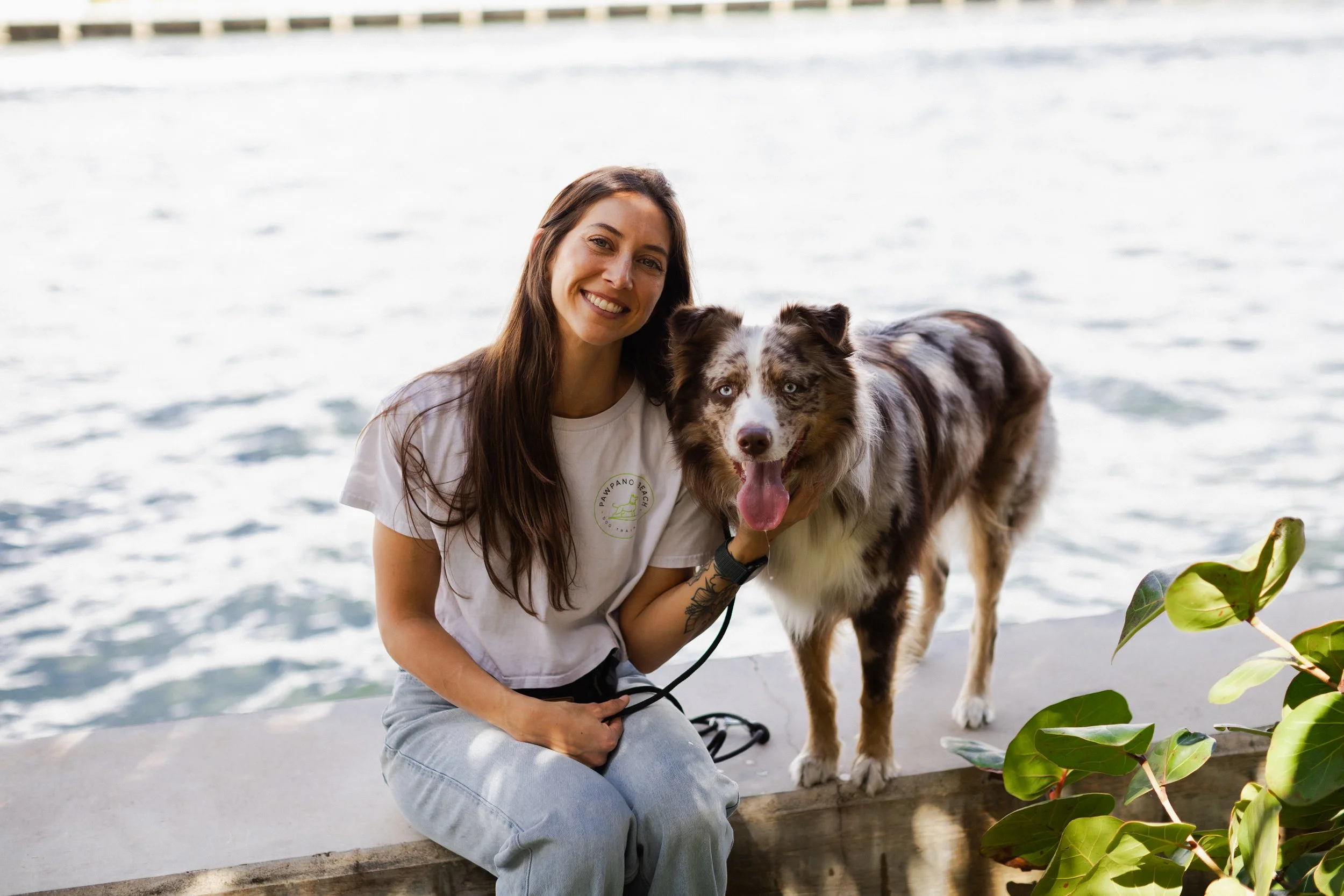 A woman with long brown hair smiling and holding a leash on her Australian Shepherd dog with blue eyes, near a body of water with gentle waves and green leaves in the foreground.