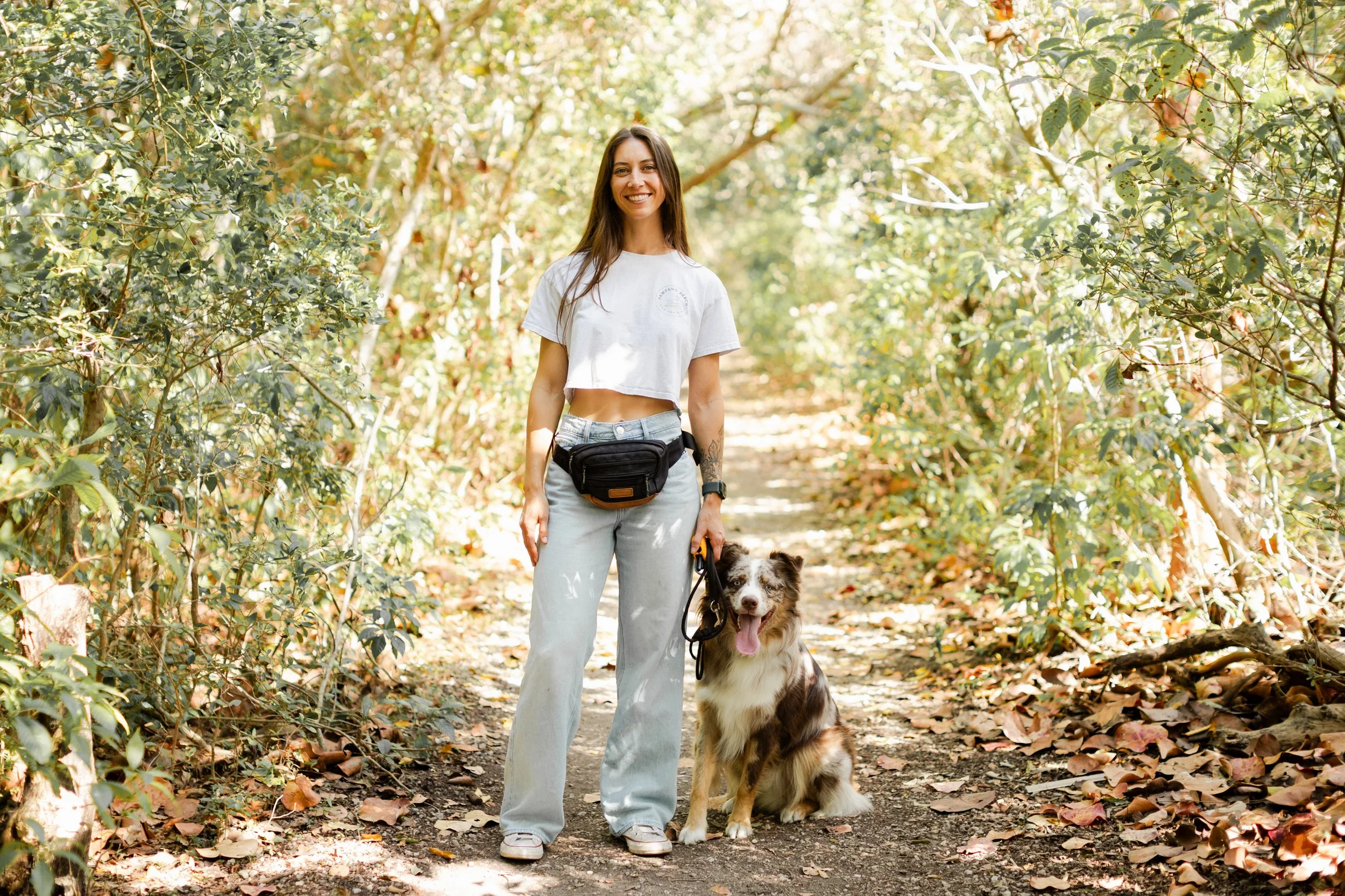 A woman smiling and standing on a wooded trail with her Australian Shepherd dog during daytime