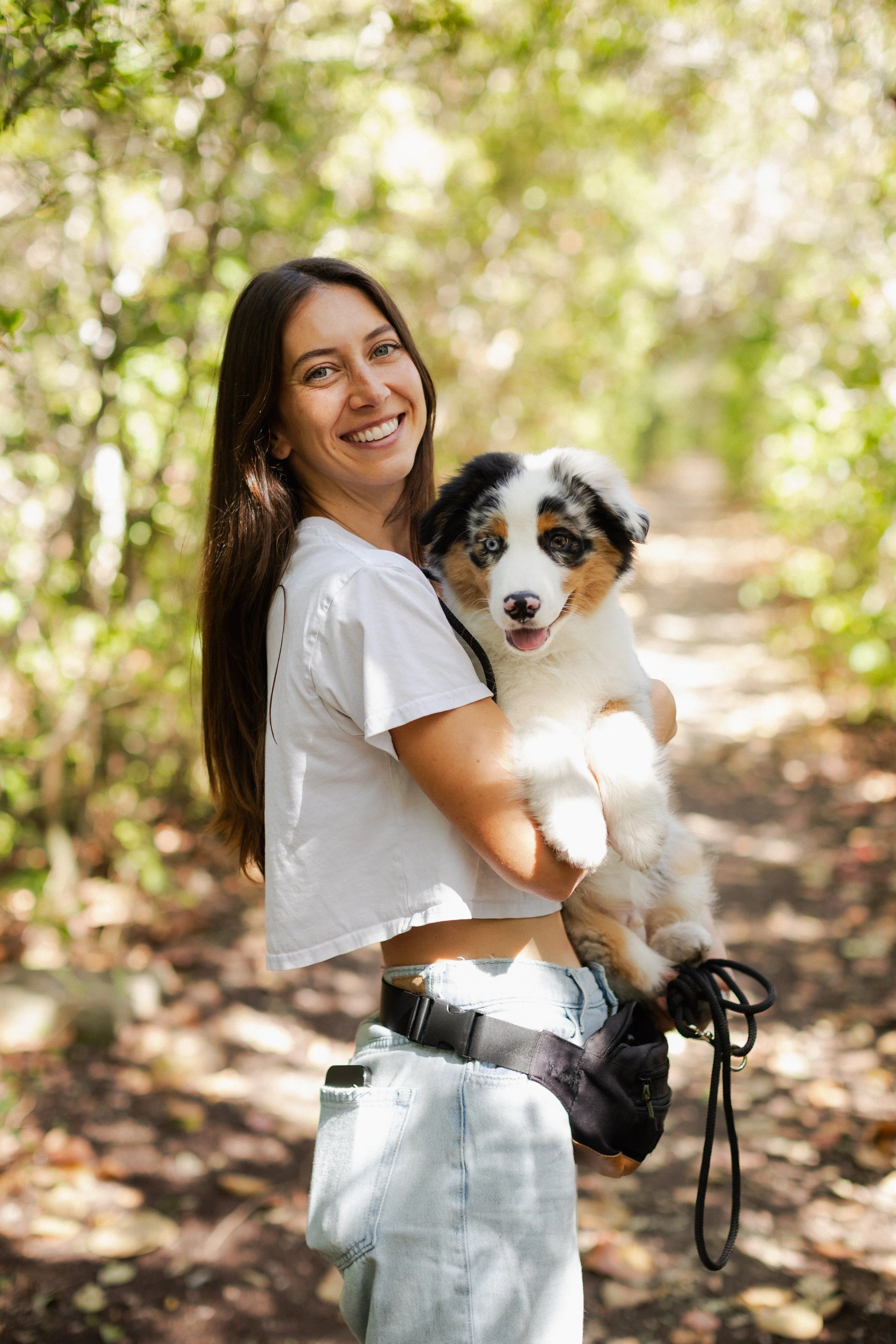 Smiling woman holding Australian Shepherd puppy outdoors on a wooded trail.