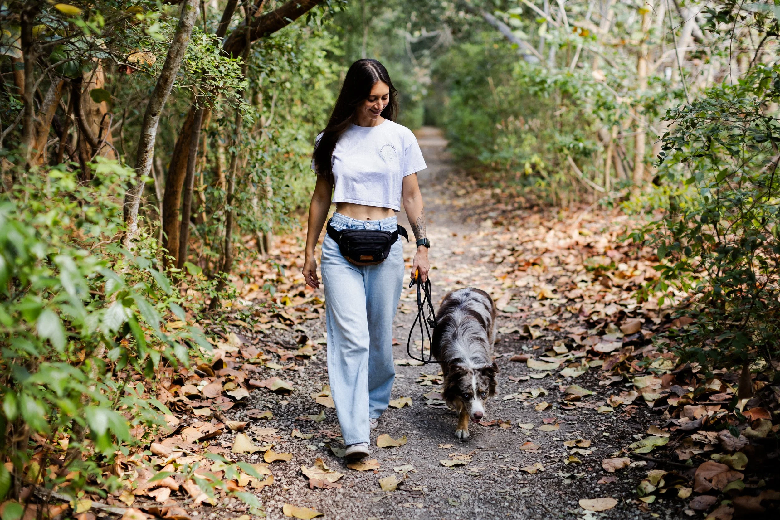 A woman walking on a forest trail with her Australian Shepherd dog, surrounded by green trees and fallen autumn leaves.