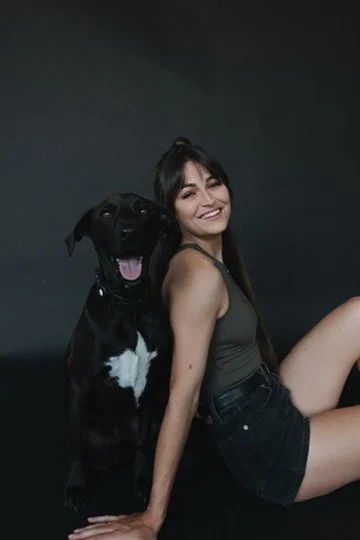 A woman with long dark hair smiling and sitting on the floor next to a large black dog with a white chest against a dark background.