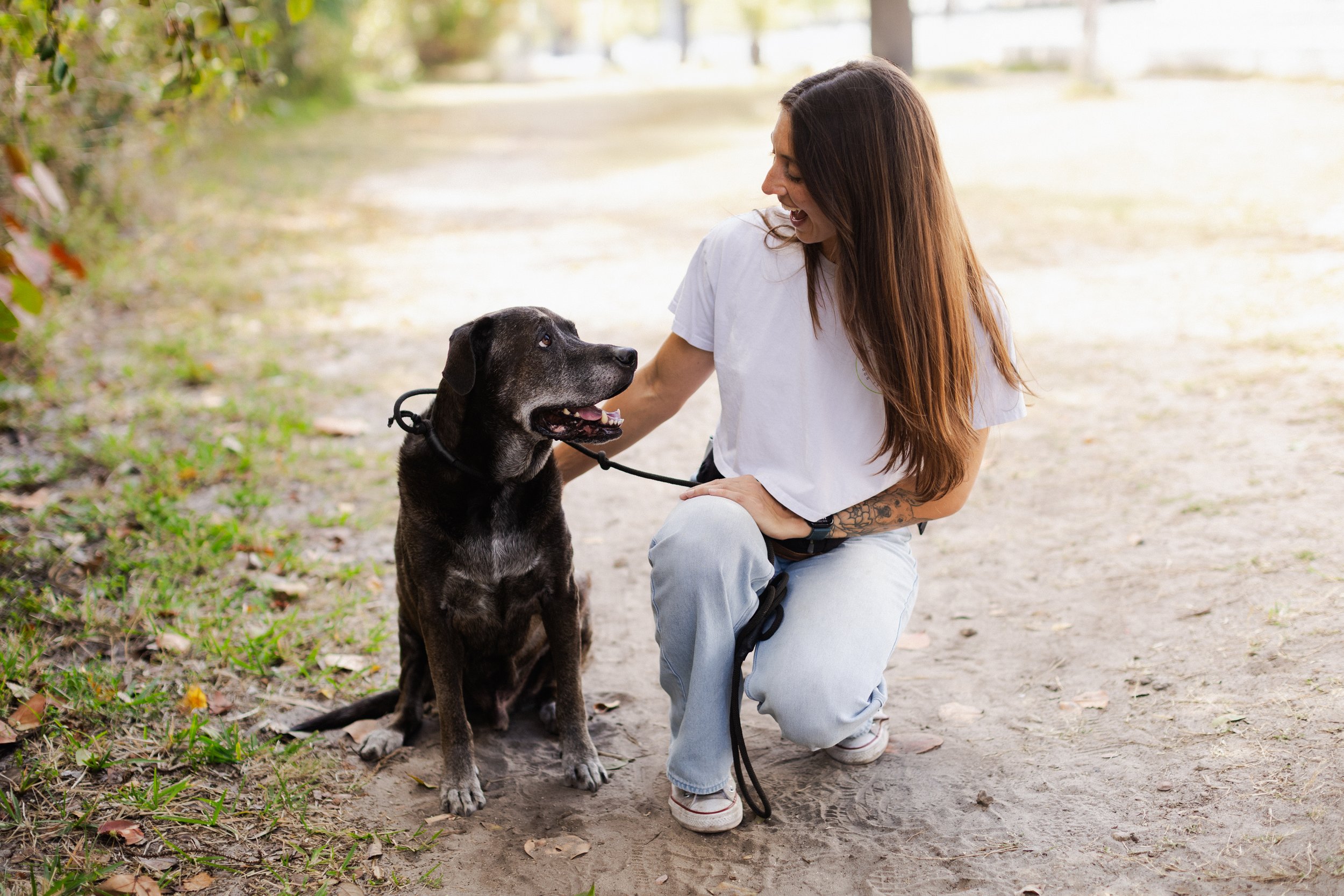 A woman with long brown hair kneeling on a dirt path, smiling at and petting a large black dog sitting beside her. The woman is wearing a white t-shirt and light blue jeans, and the dog is wearing a collar and leash, surrounded by a park with trees and greenery in the background.