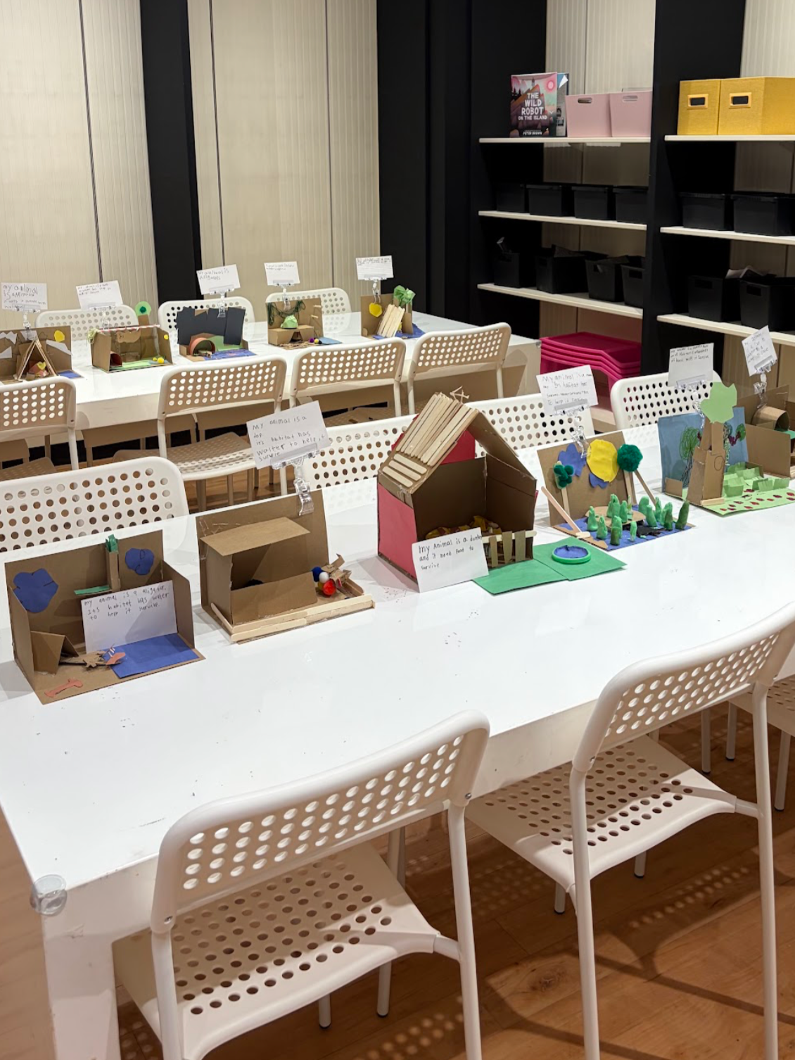 Display of small animal dioramas and craft projects on a white table in a classroom or library setting, with storage shelves in the background.