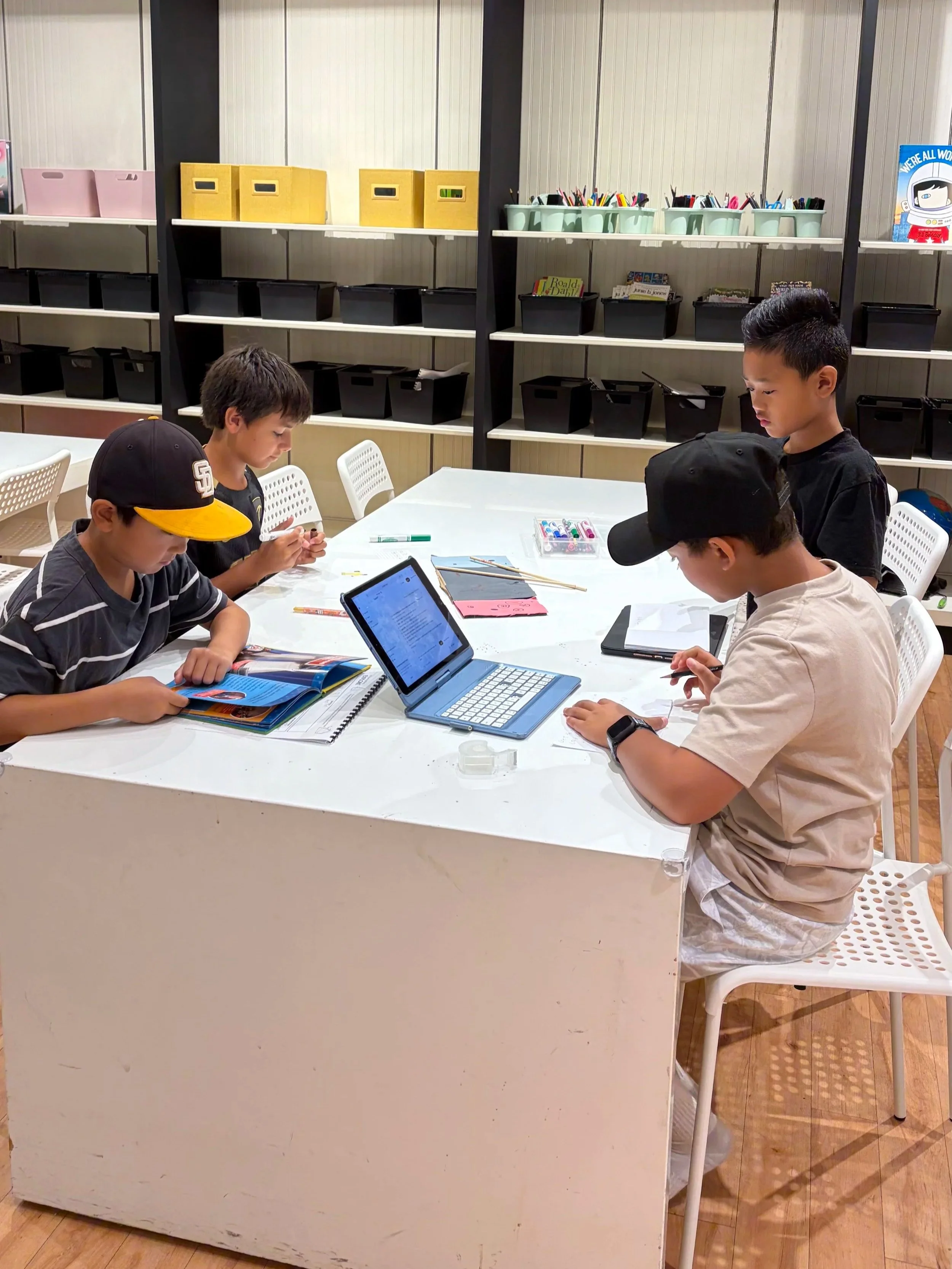 Four children sitting at a white table in a classroom, engaged in activities using tablets, books, and stationery, with a background of shelves filled with colorful bins and books.
