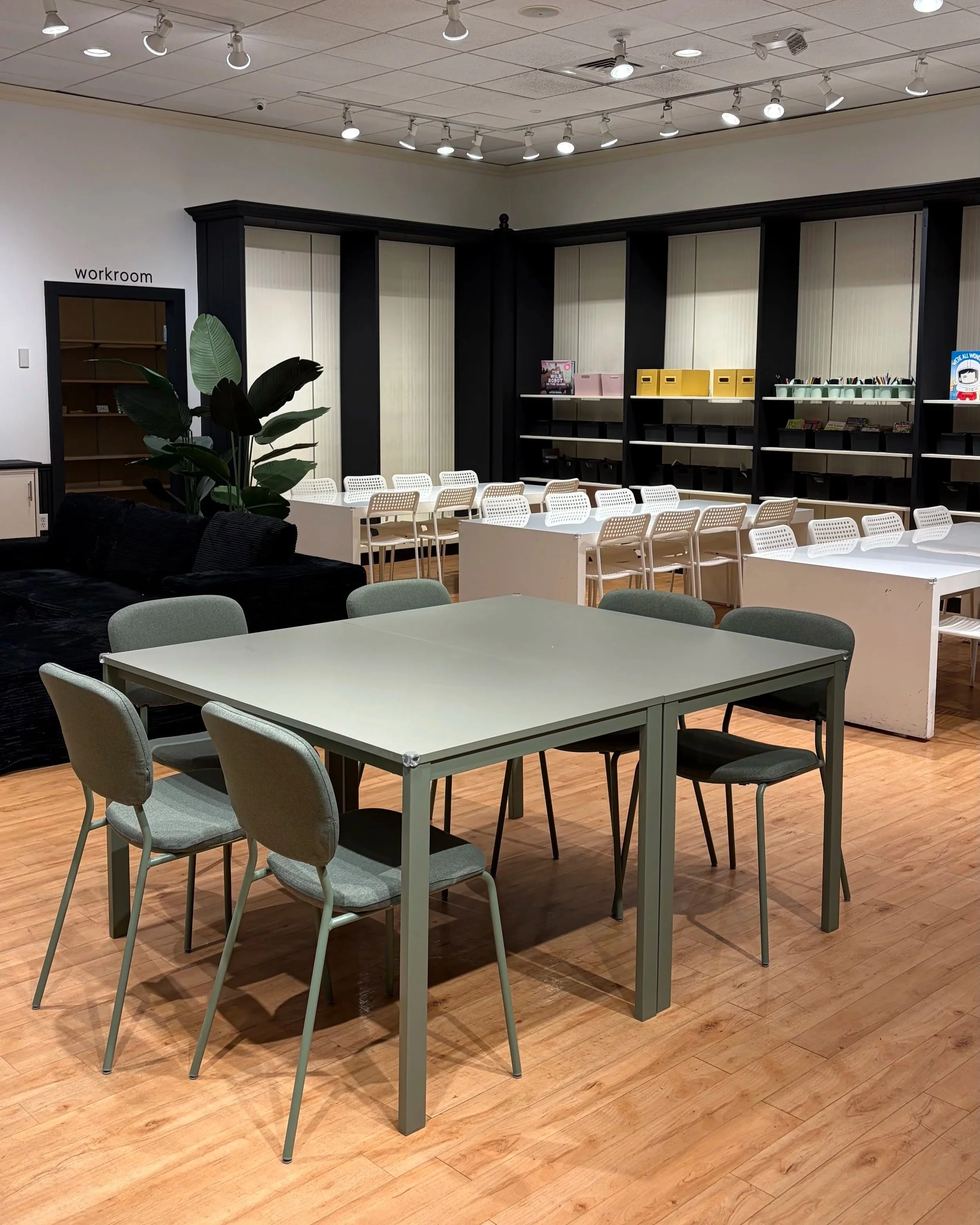 A conference room with white tables and gray chairs, black shelving units along the wall, a large plant with broad green leaves in the corner, and a white ceiling with multiple track lights. There are some books and storage boxes on the shelves, and a door labeled 'workroom' on the side.