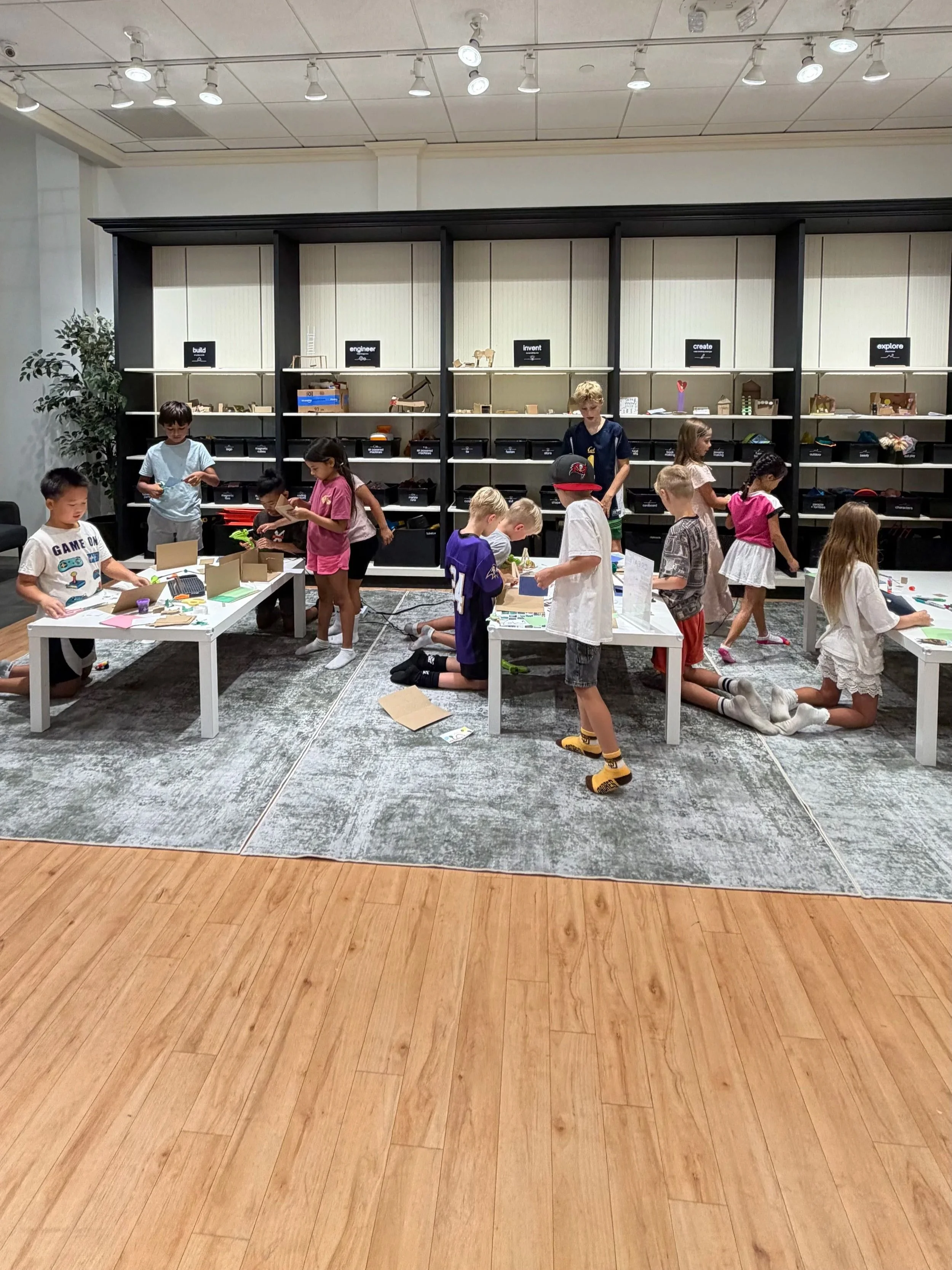 Children participating in a creative workshop in an indoor space with shelves labeled such as build, engineer, invent, create, explore, and a wooden floor and ceiling lights.