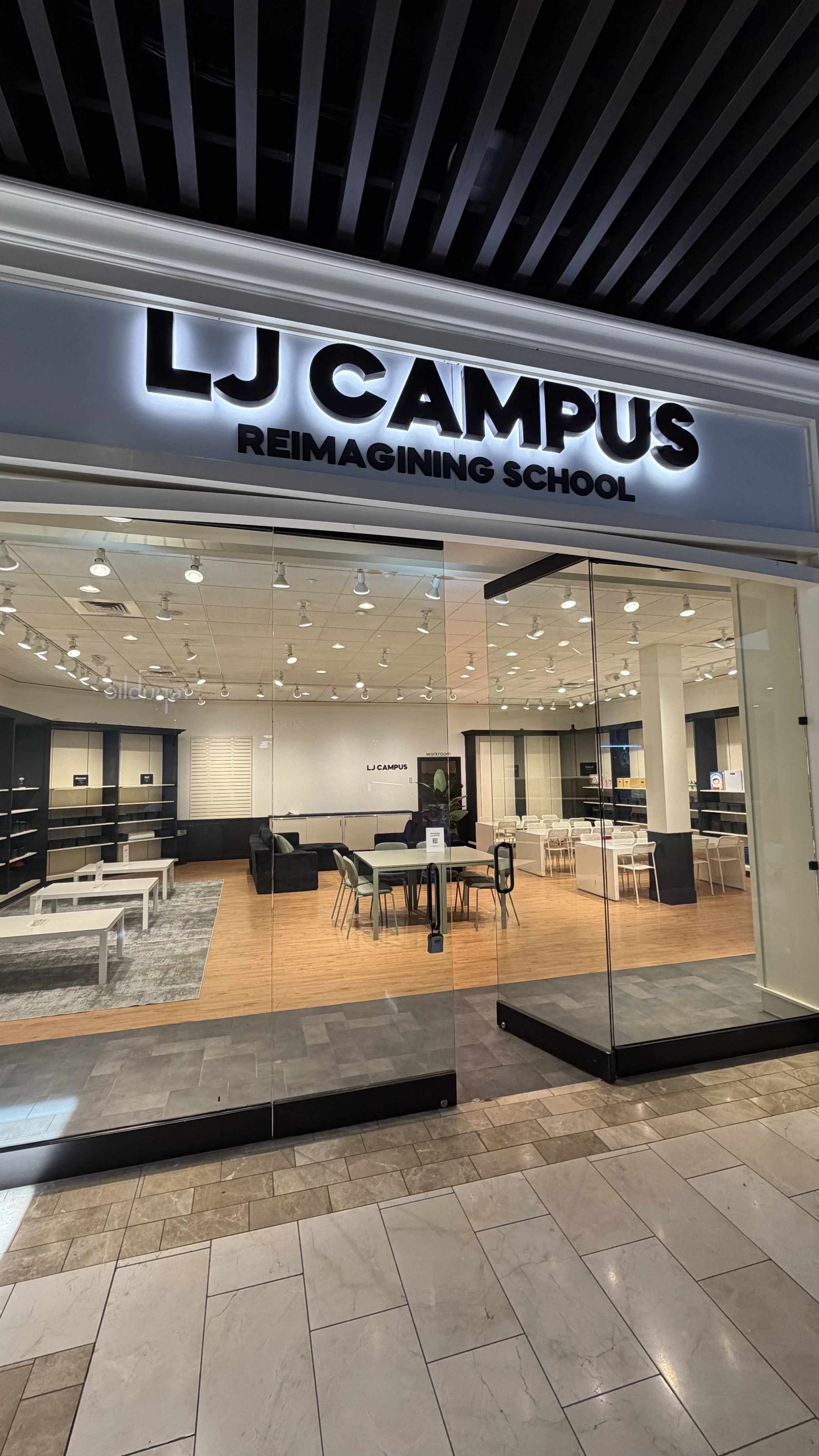 storefront with illuminated sign reading 'LJ CAMPUS REIMAGINING SCHOOL' and large glass windows showing a modern, empty interior with tables, chairs, and bookshelves.