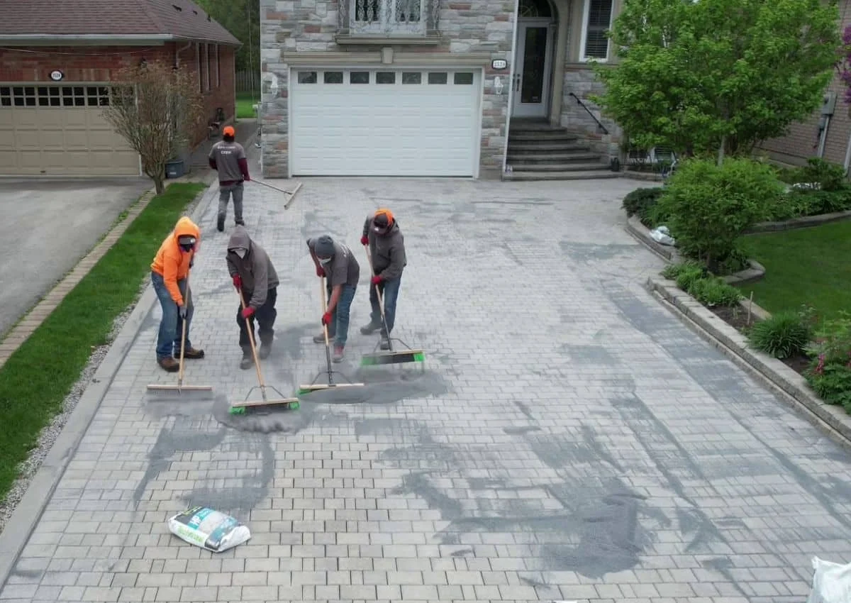 Five workers are paving a driveway with bricks in front of a house, using tools to spread and level the pavement.