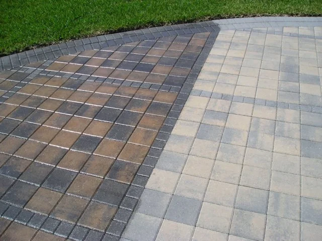Paved driveway with gray and brown interlocking bricks, bordered by a strip of darker bricks and a grassy lawn.