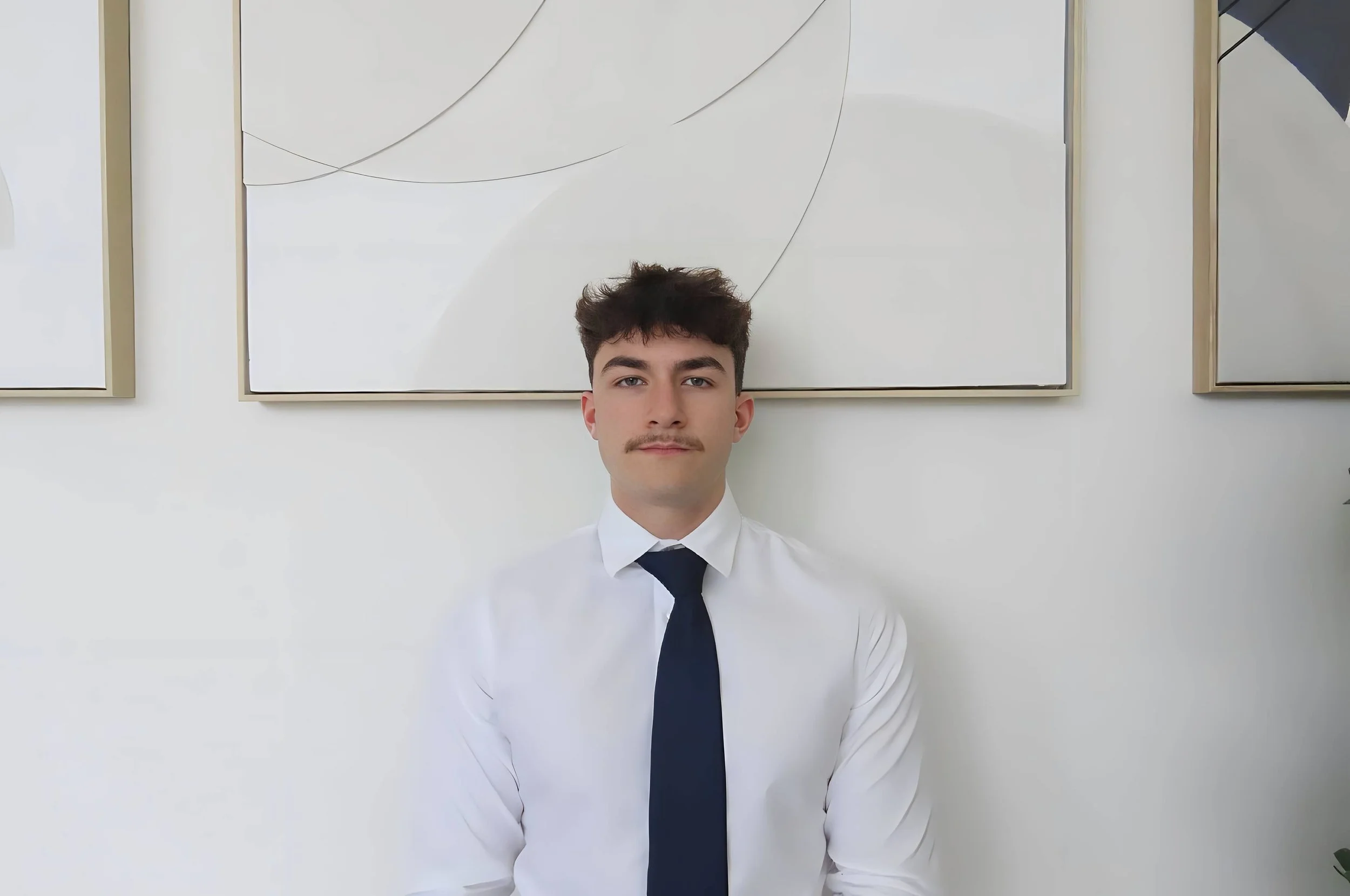 A woman with short blonde hair sitting on a beige bench, writing in a notebook with sunlight casting shadows from a window and a large leafy plant beside her.