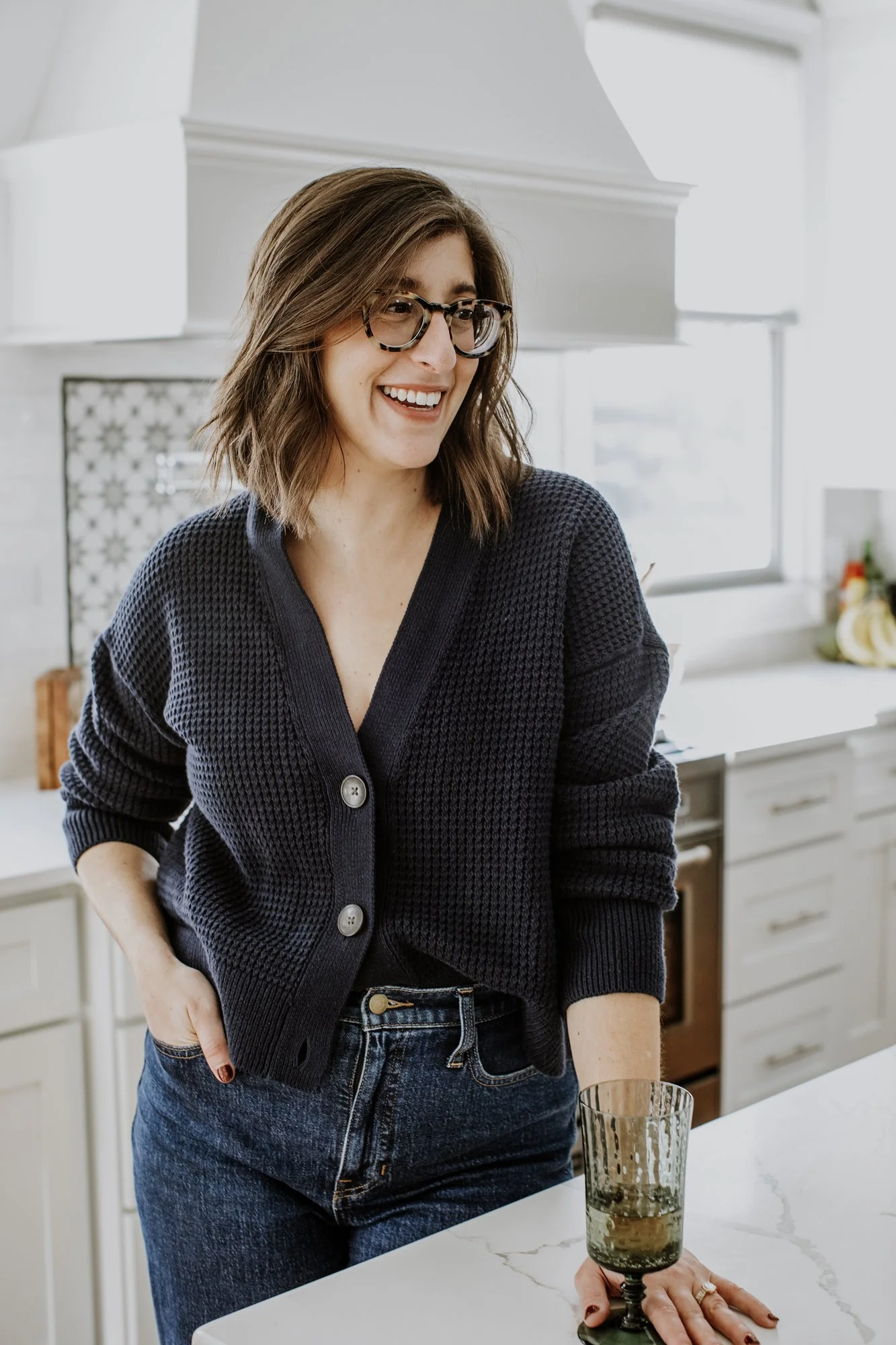 A woman with shoulder-length brown hair, wearing glasses and a dark navy cardigan, smiling in a bright kitchen with white cabinets and a marble countertop.