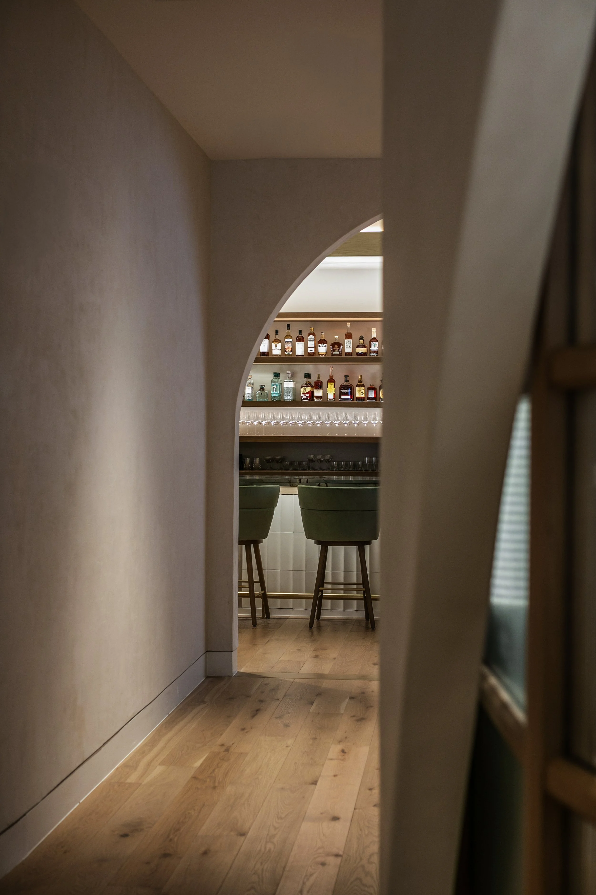 View into a bar area through a narrow passage with an arch, showing a backlit shelf of various liquor bottles and a row of glasses, with green bar stools in front.