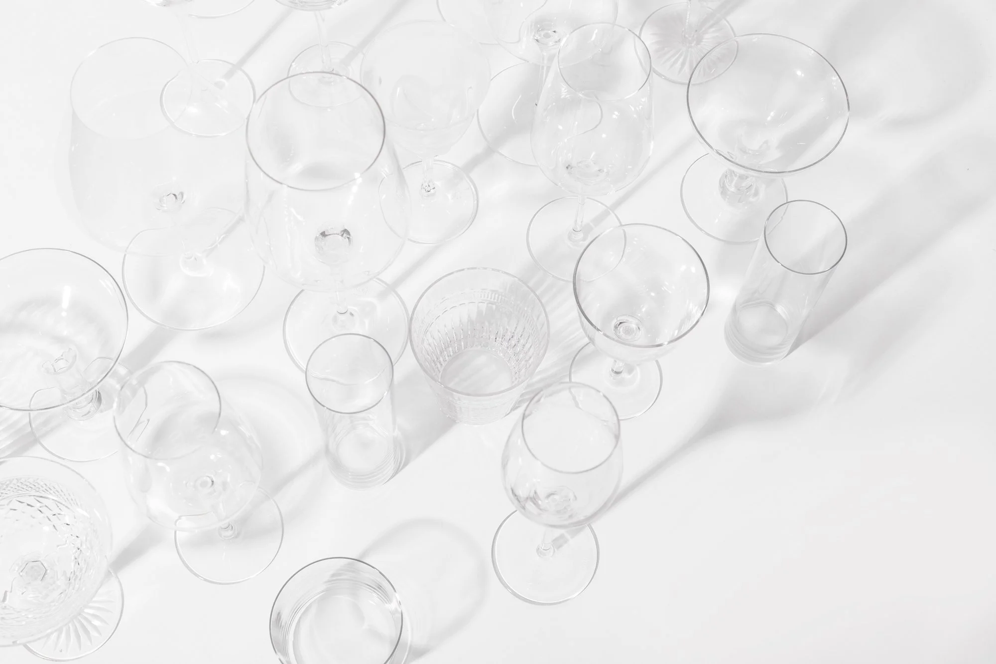 Various empty transparent glassware, including wine glasses and tumblers, arranged on a white surface, with shadows cast by the glasses.