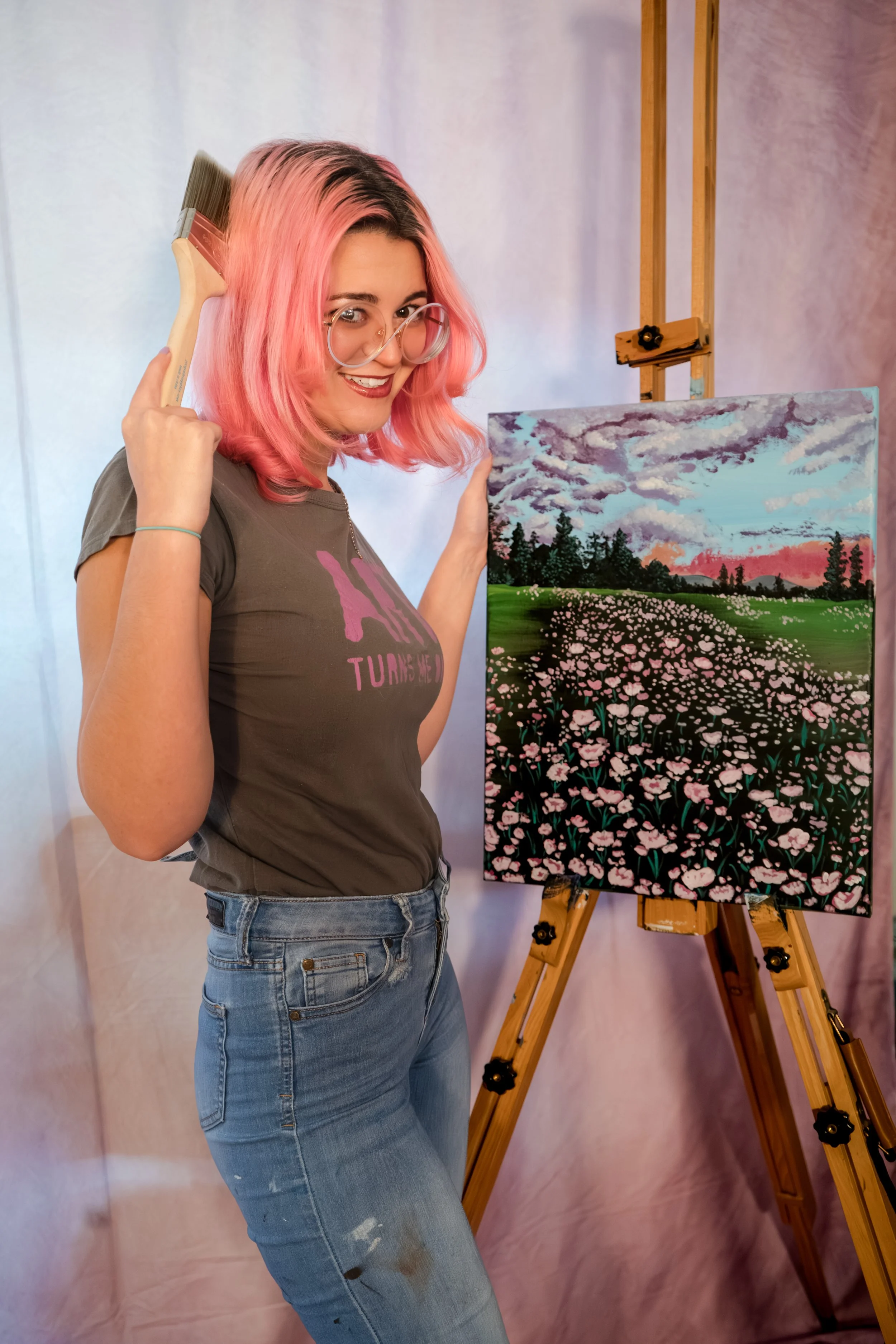 A woman with pink hair and glasses holding a paintbrush behind her head, standing next to an easel with a landscape painting of a field of pink flowers under a cloudy sky.