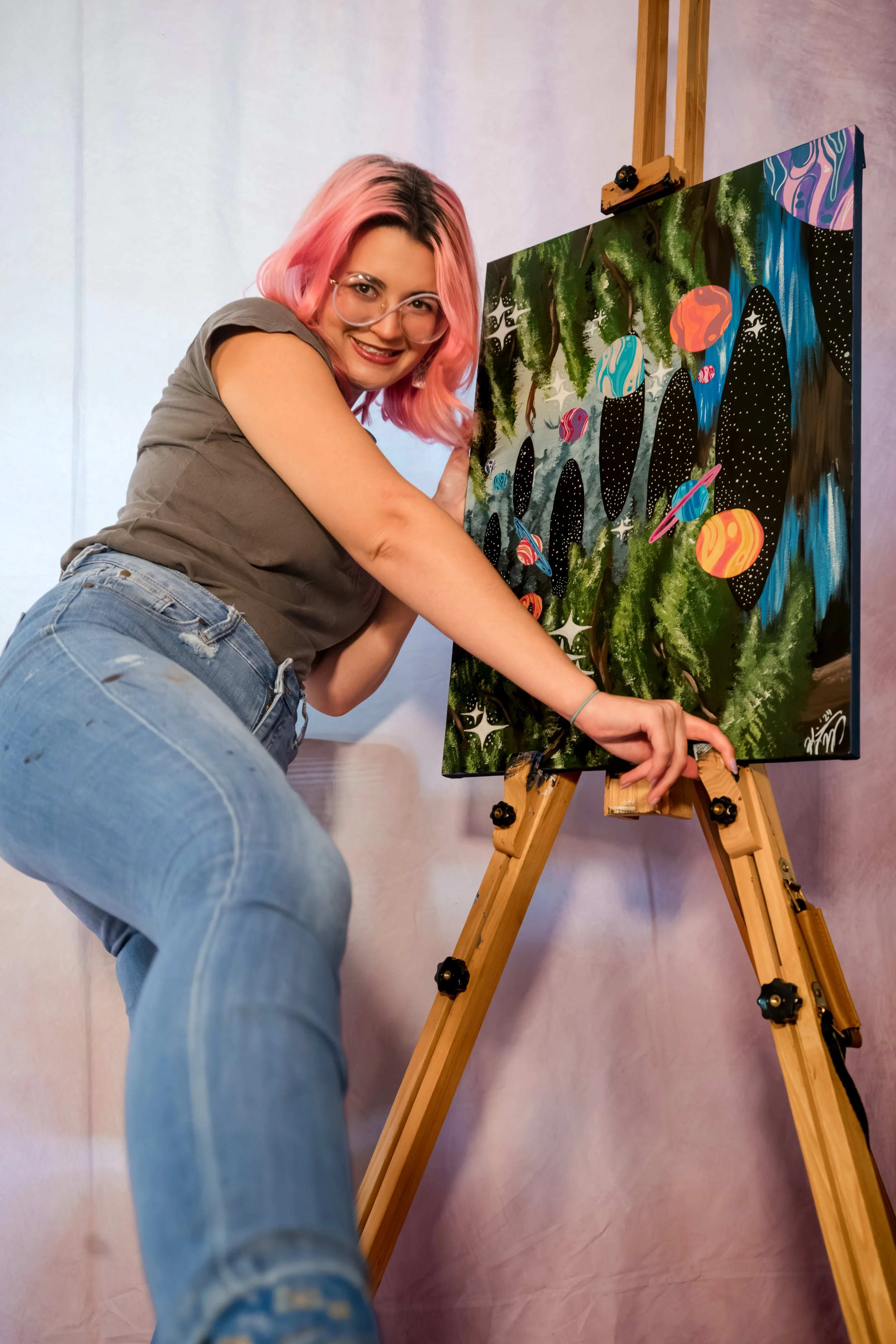 A woman with pink hair, glasses, and a gray shirt is painting a colorful galaxy on a canvas on an easel.