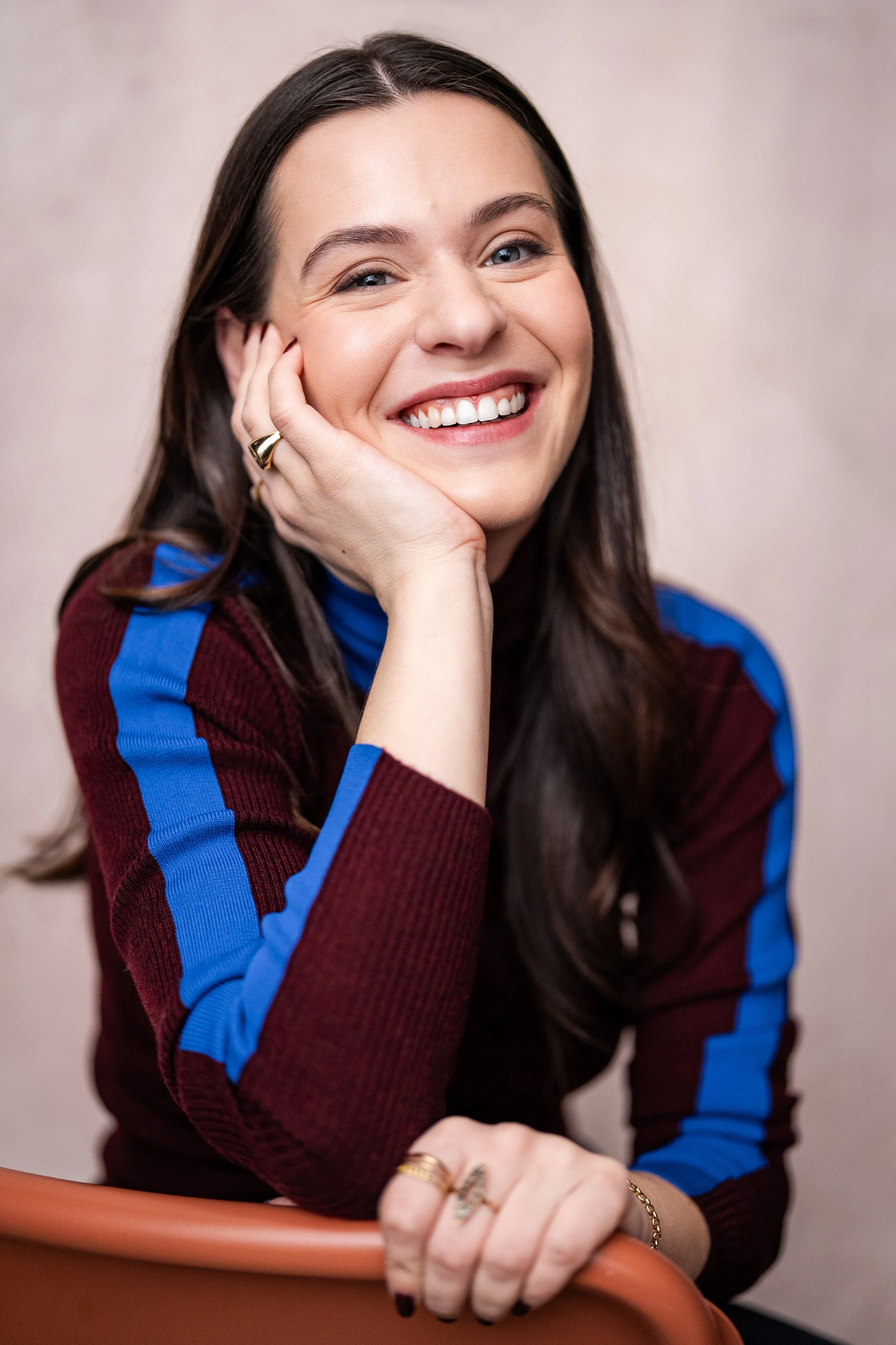 A woman with long brown hair wearing a maroon sweater with blue stripes, smiling with her hand resting on her chin, showing her teeth.