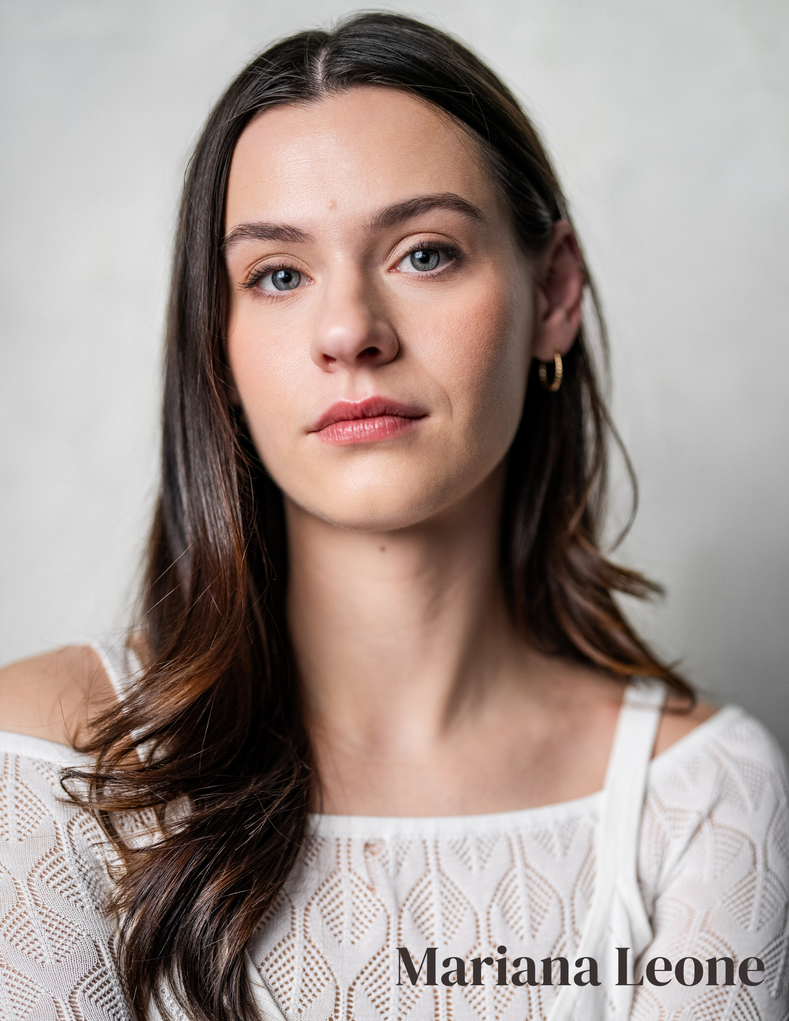 A portrait of a young woman with long brown hair, blue eyes, and fair skin, wearing a white patterned top and gold hoop earrings, against a plain background. The name Mariana Leone is written at the bottom right corner of the image.