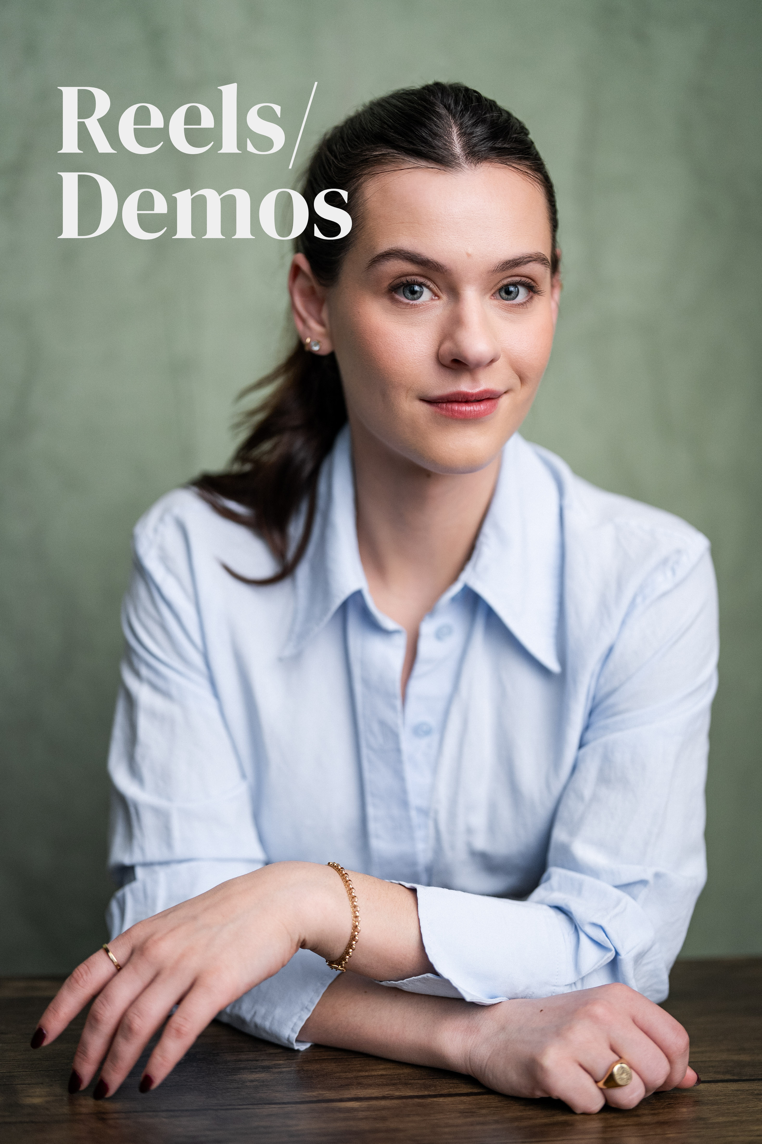 A young woman with dark hair pulled back, wearing a light blue button-up shirt, sitting at a wooden table with a green background. The text 'Reels/Demos' appears in the upper left corner.