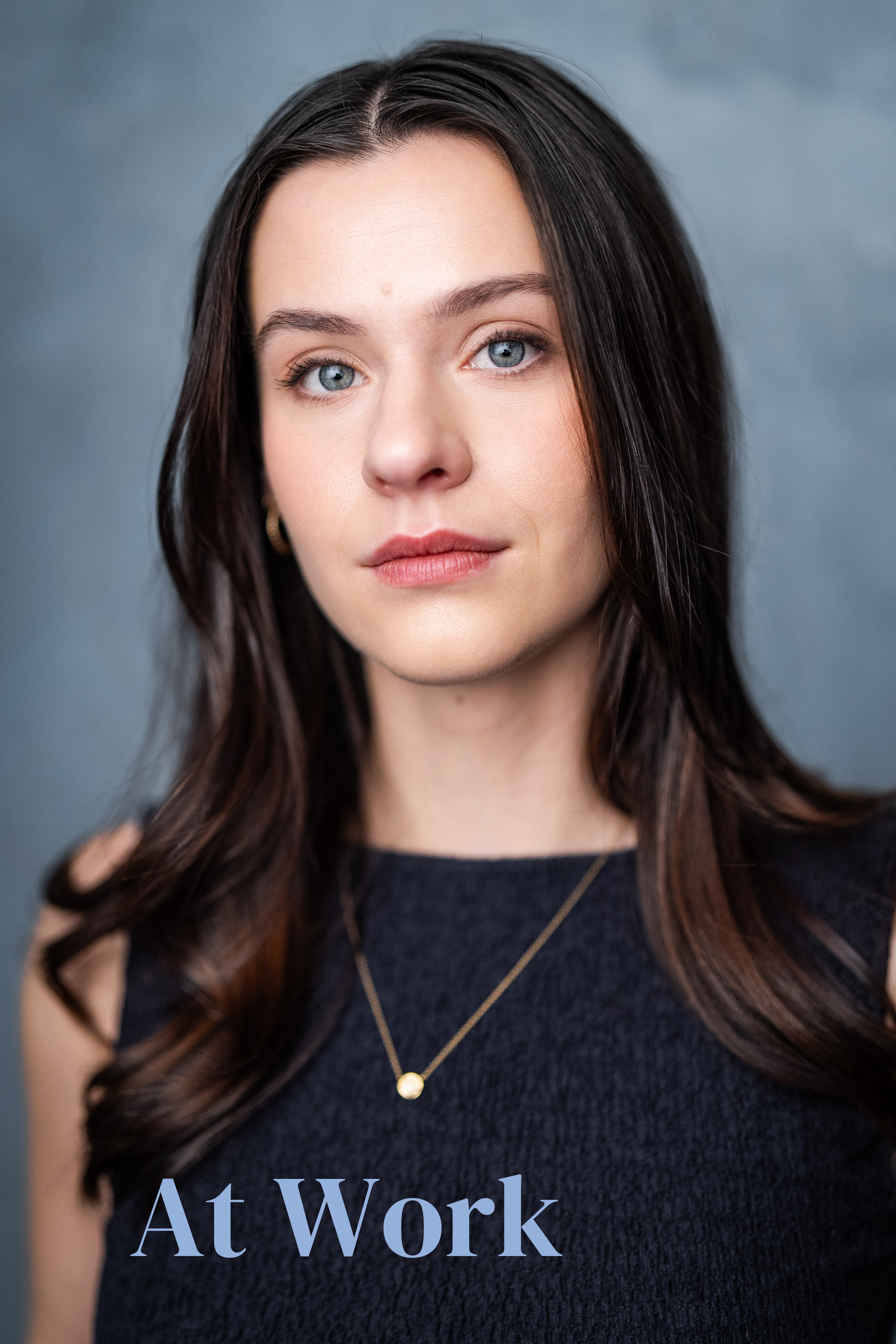 A young woman with dark brown hair, blue eyes, and a neutral expression, wearing a navy sleeveless top and a delicate gold necklace, against a gray background. Text overlay says 'At Work'.