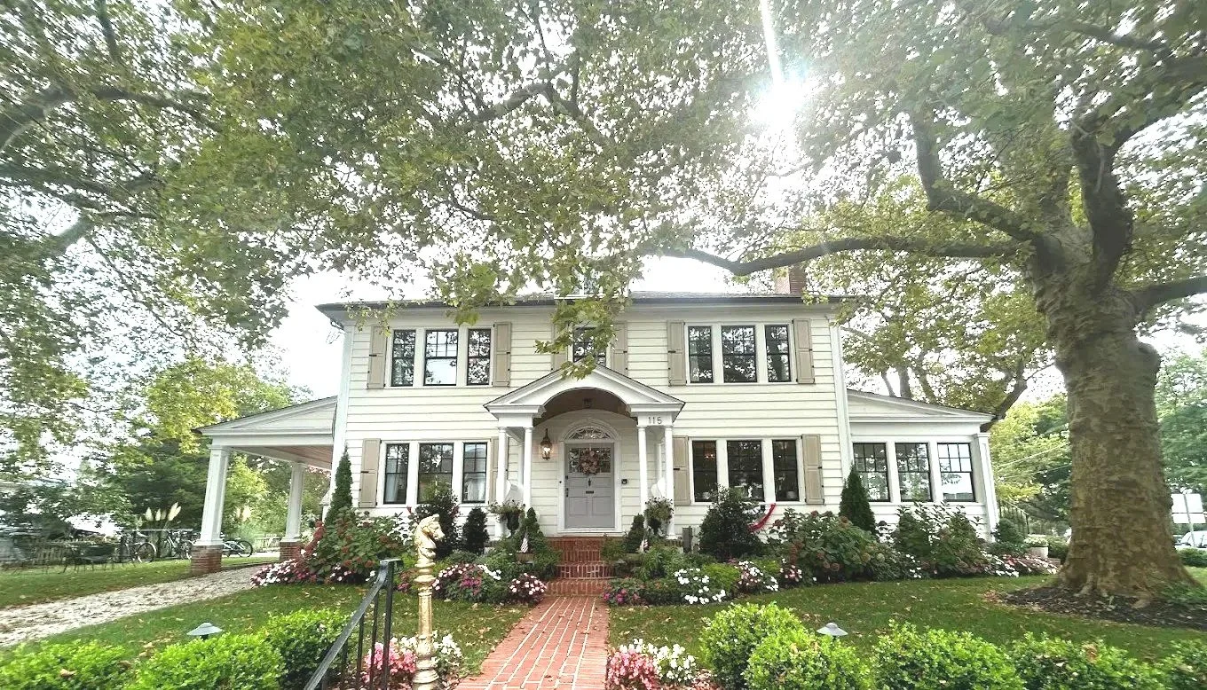 White two-story house with a porch, surrounded by a manicured lawn and colorful flowers, large trees, and a bright sky.