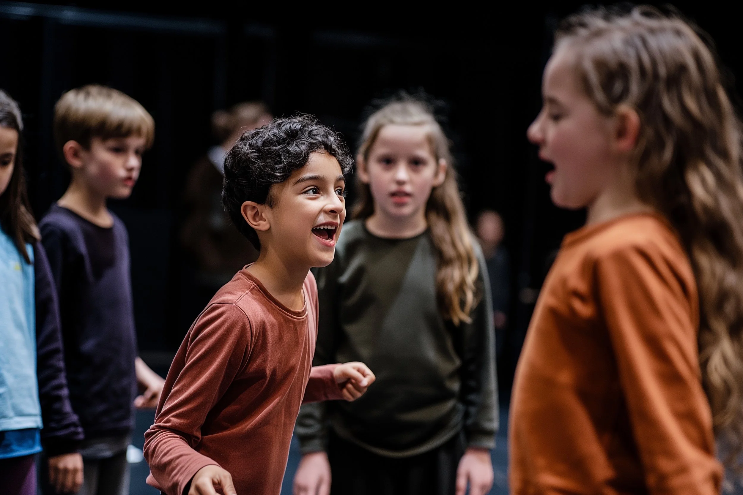 A group of primary school children taking part in a drama class. In the foreground, a boy with curly dark hair laughs with delight mid-scene, while other children stand around him in various states of concentration and performance.