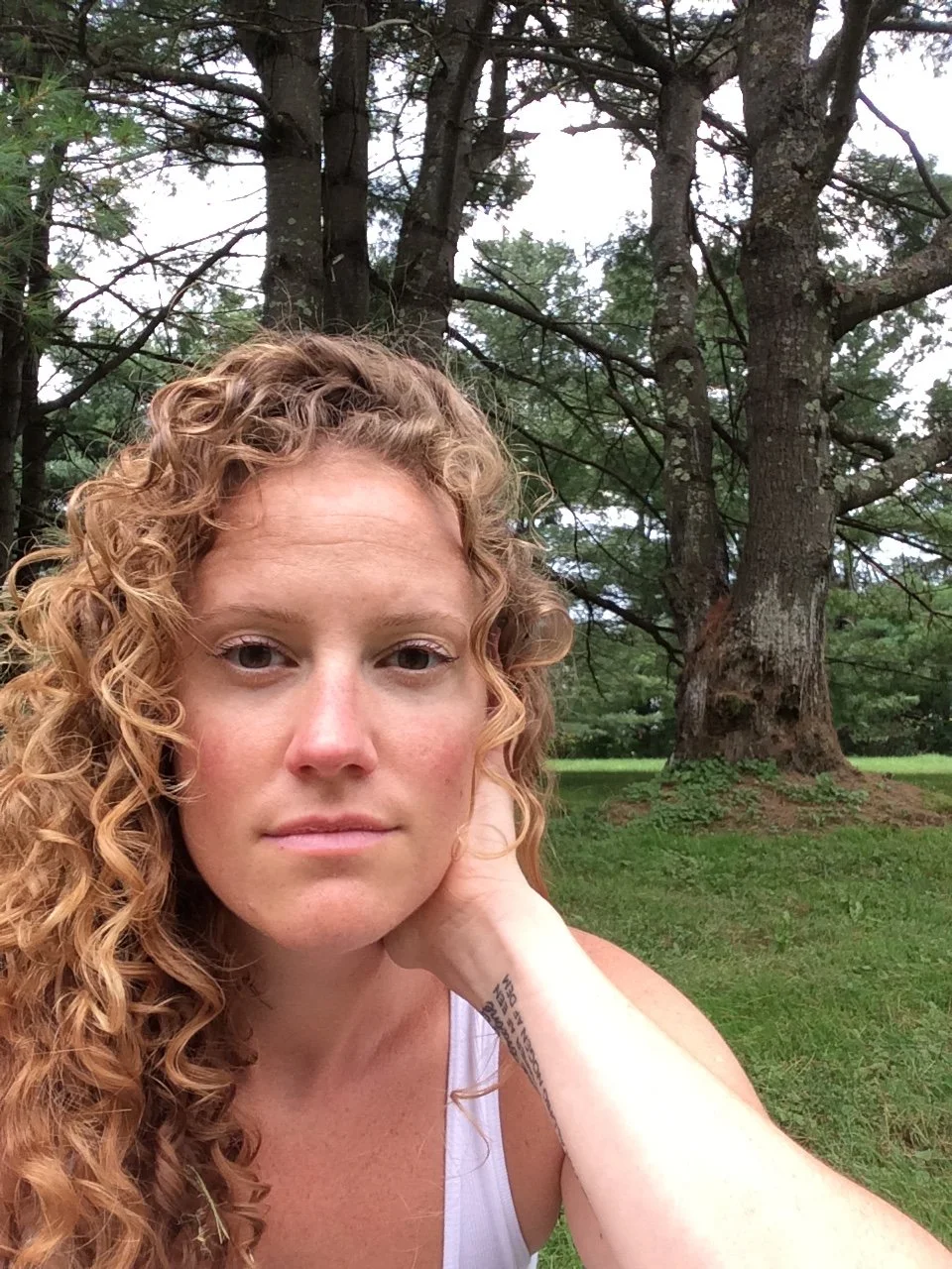 A woman with curly red hair taking a selfie outdoors in front of a large tree with green leaves.