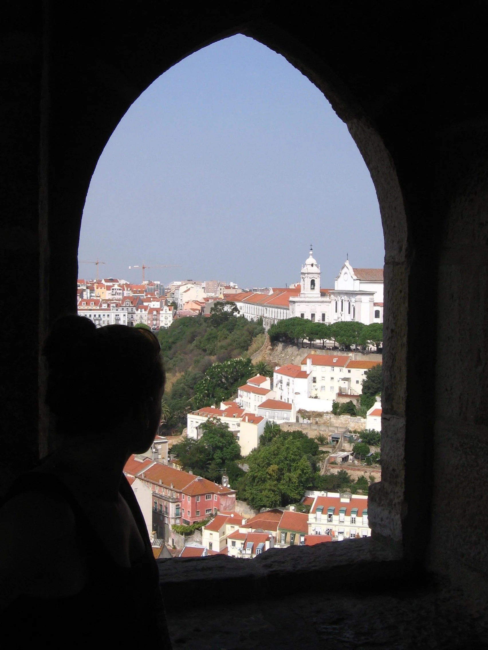 A view of a cityscape through a small, arched window in a dark stone wall, showing white buildings with red-tiled roofs, a church with a bell tower, and cranes in the distance.