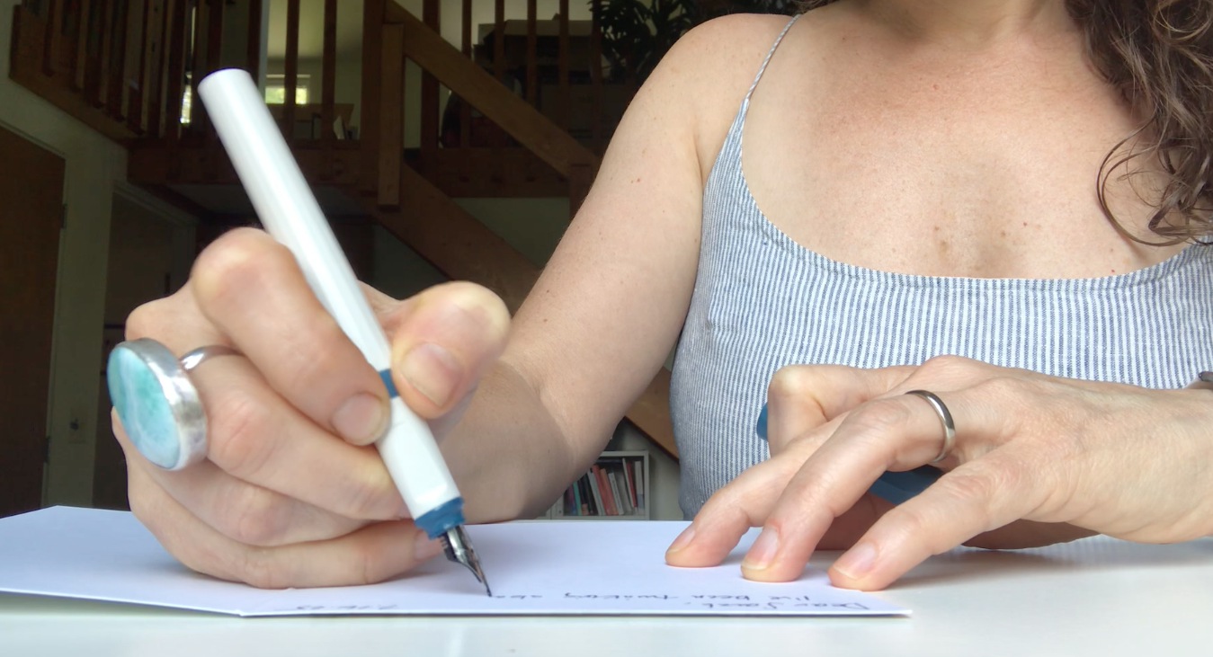 A woman wearing a striped tank top is sitting at a table, writing with a white pen on a sheet of paper.