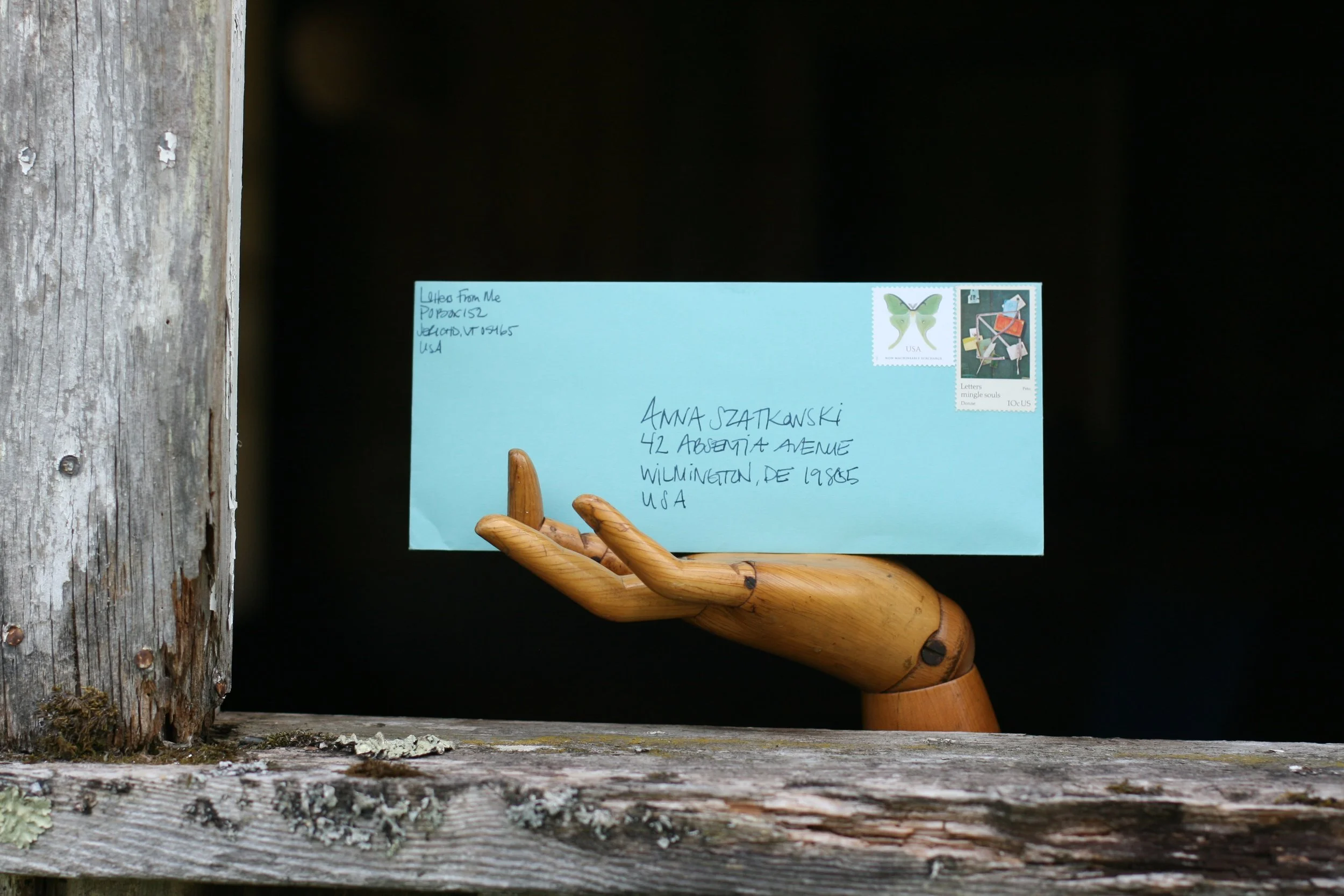 A light blue envelope with handwritten address and two postage stamps, one with a butterfly and the other with abstract art, resting on a wooden hand sculpture placed on a weathered wooden surface near a wooden post.