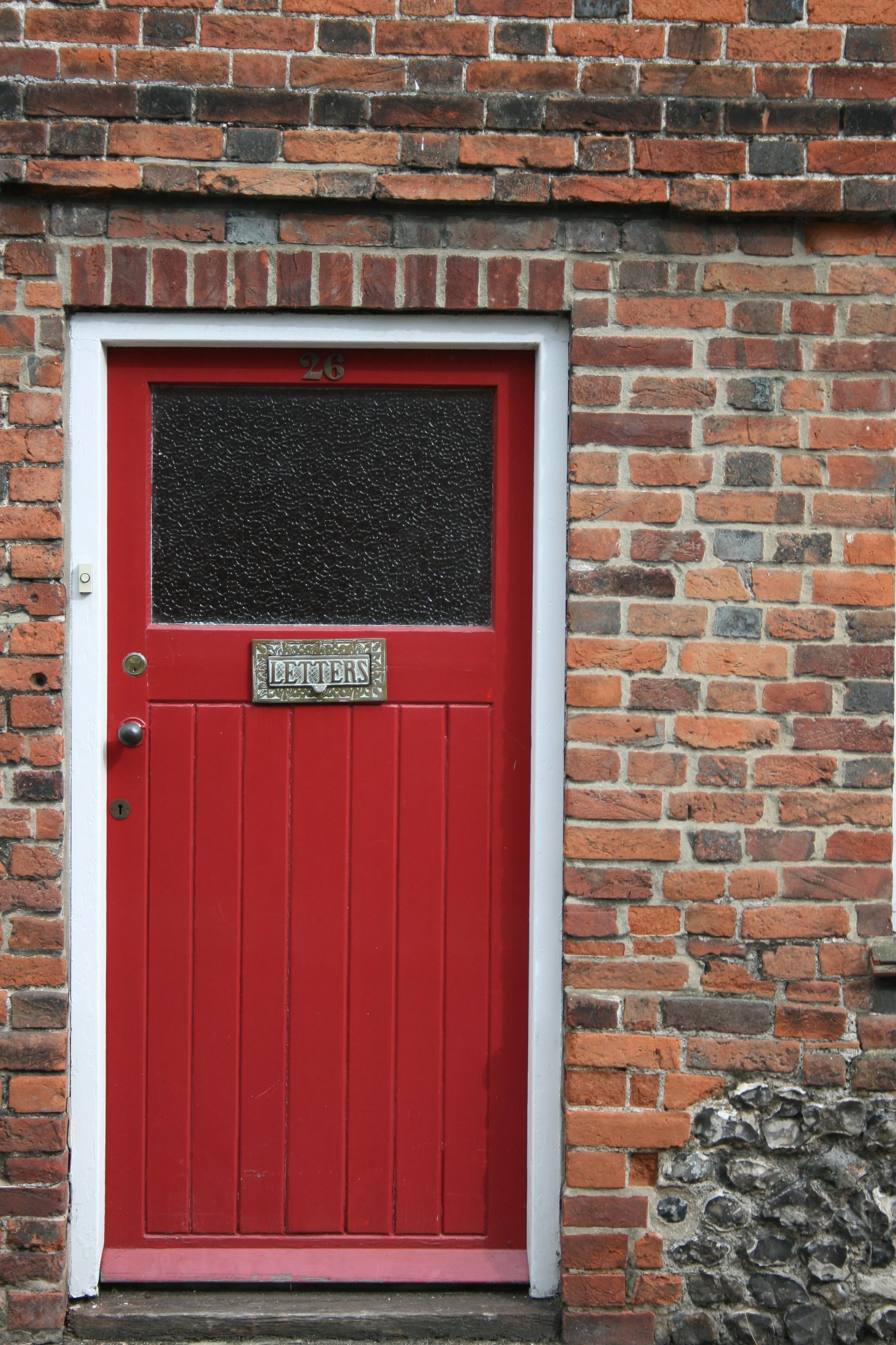 A red door in a brick wall with a textured glass window, a letterbox with the word 'LETTERS' on it, and the number 26 above the window.