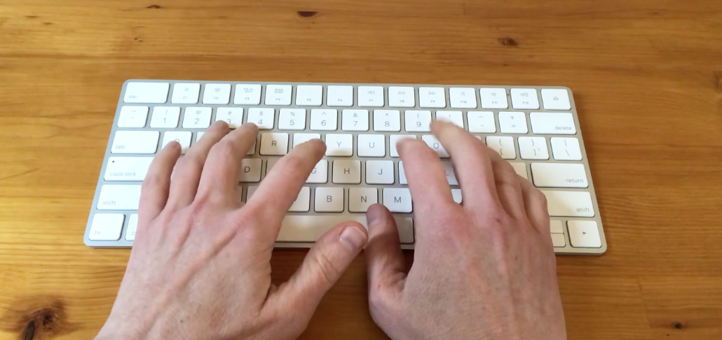 Two hands typing on a white computer keyboard on a wooden desk.
