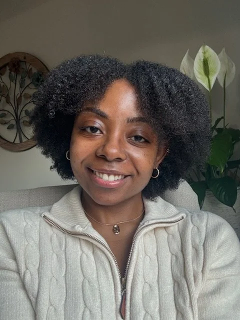 A young woman with natural curly hair smiling at the camera, wearing a light-colored zip-up sweater and small hoops earrings, with a plant and wall decoration in the background.