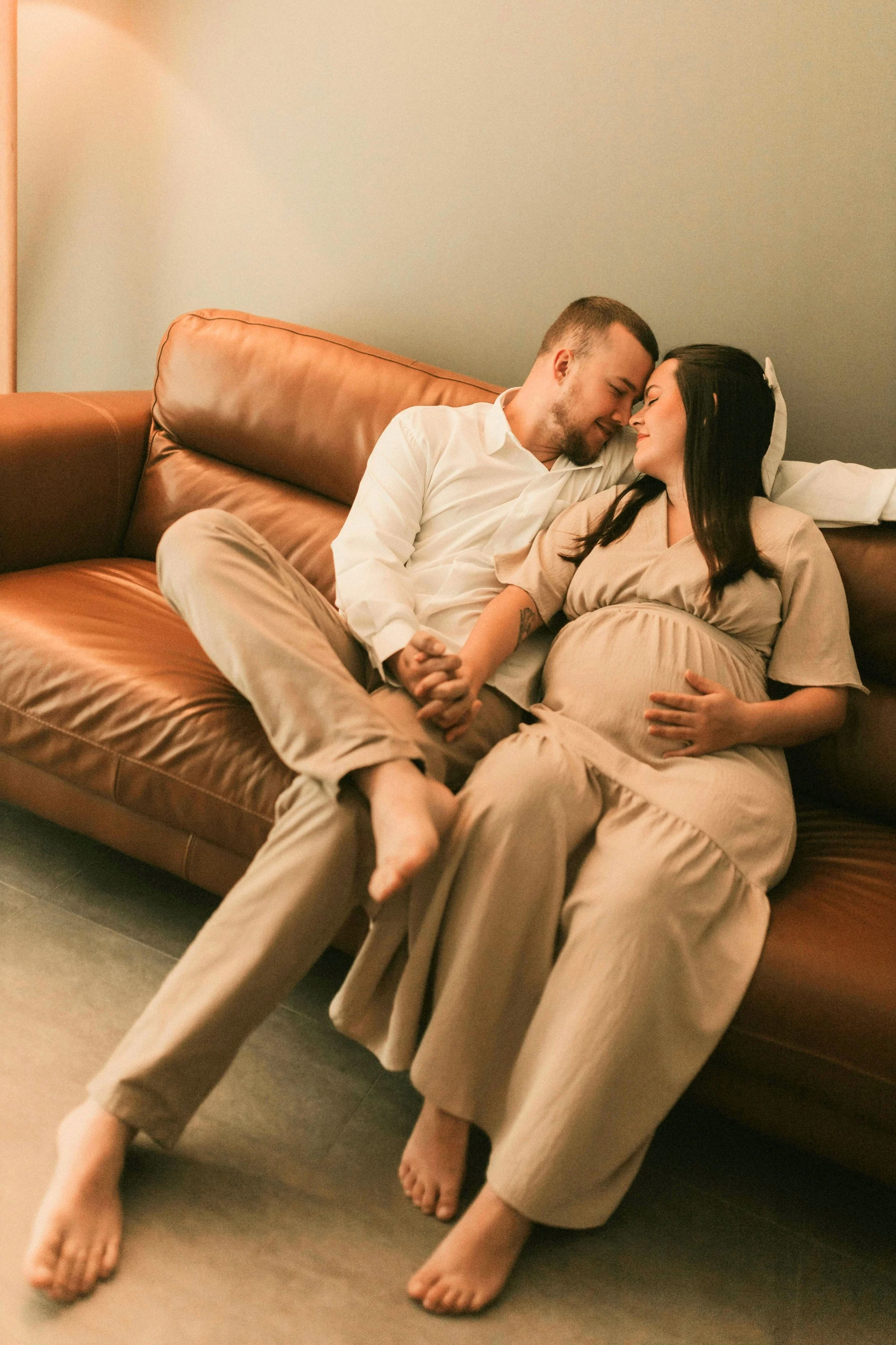 A couple sitting on a leather couch, with the woman visibly pregnant, leaning against each other with eyes closed, holding hands, in a cozy room.