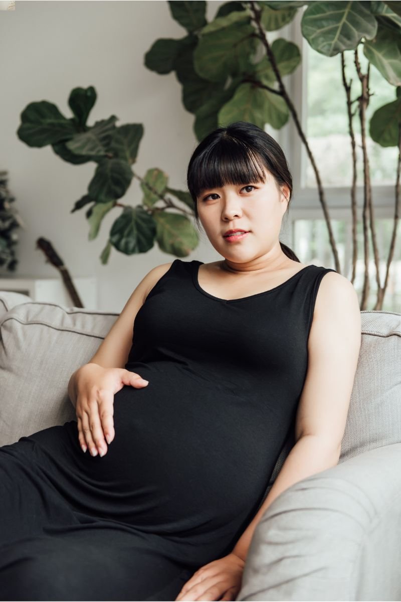 A pregnant woman sitting on a sofa in a living room with large green plants in the background, wearing a black sleeveless dress.