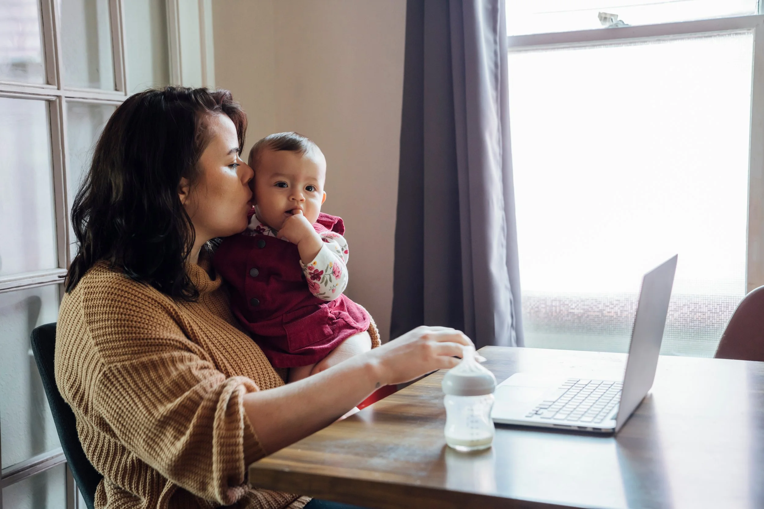 A woman with dark, wavy hair, wearing a brown knitted sweater, is holding a baby girl with short light hair and wearing a red vest with floral sleeves. They are sitting at a wooden table in front of a laptop, with a window and curtains in the background. The woman appears to be working or browsing on the laptop while holding the baby, who has fingers in her mouth.