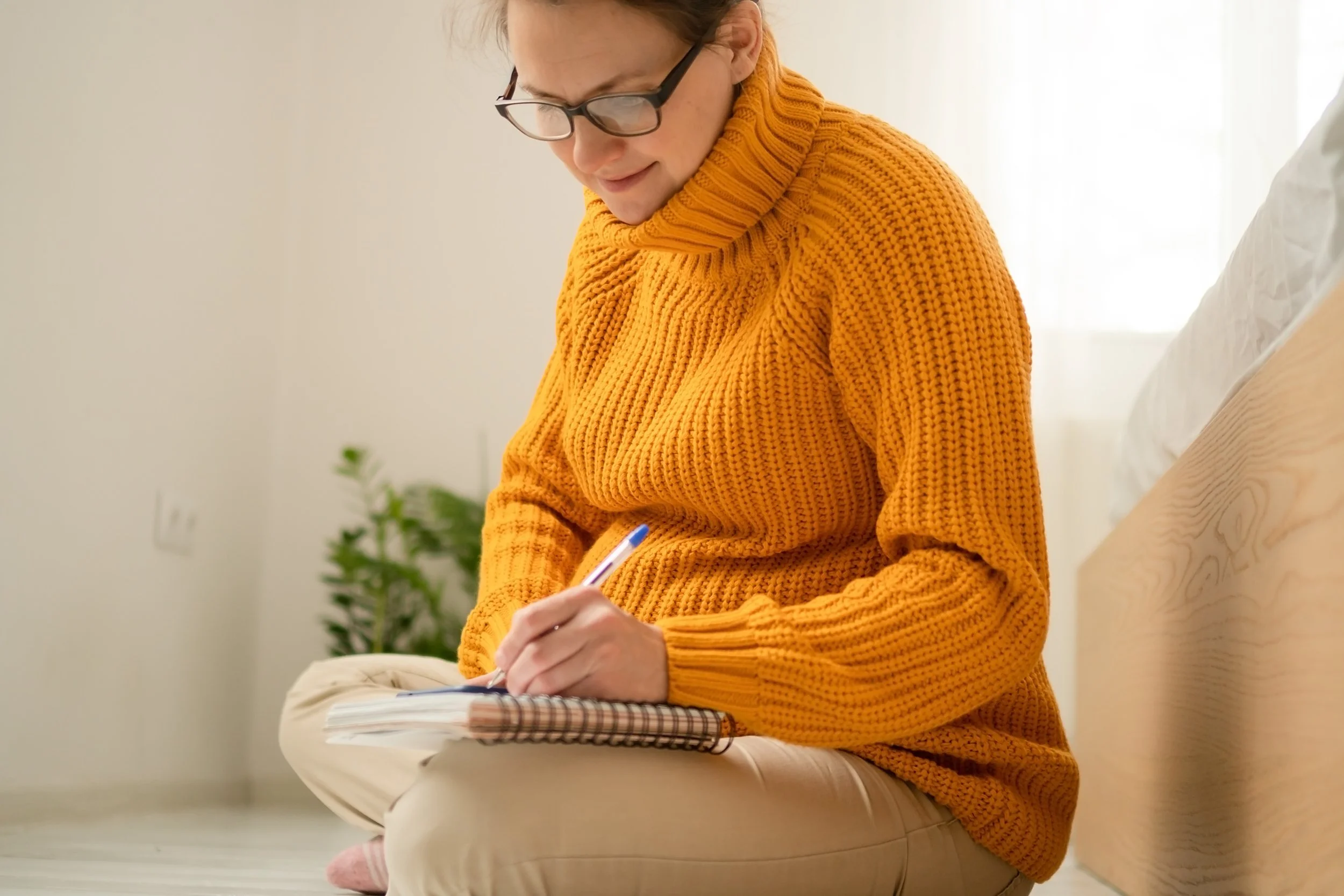 A pregnant woman with glasses and reddish hair, wearing an orange chunky knitted sweater, is sitting cross-legged on the floor and journaling in a notebook.