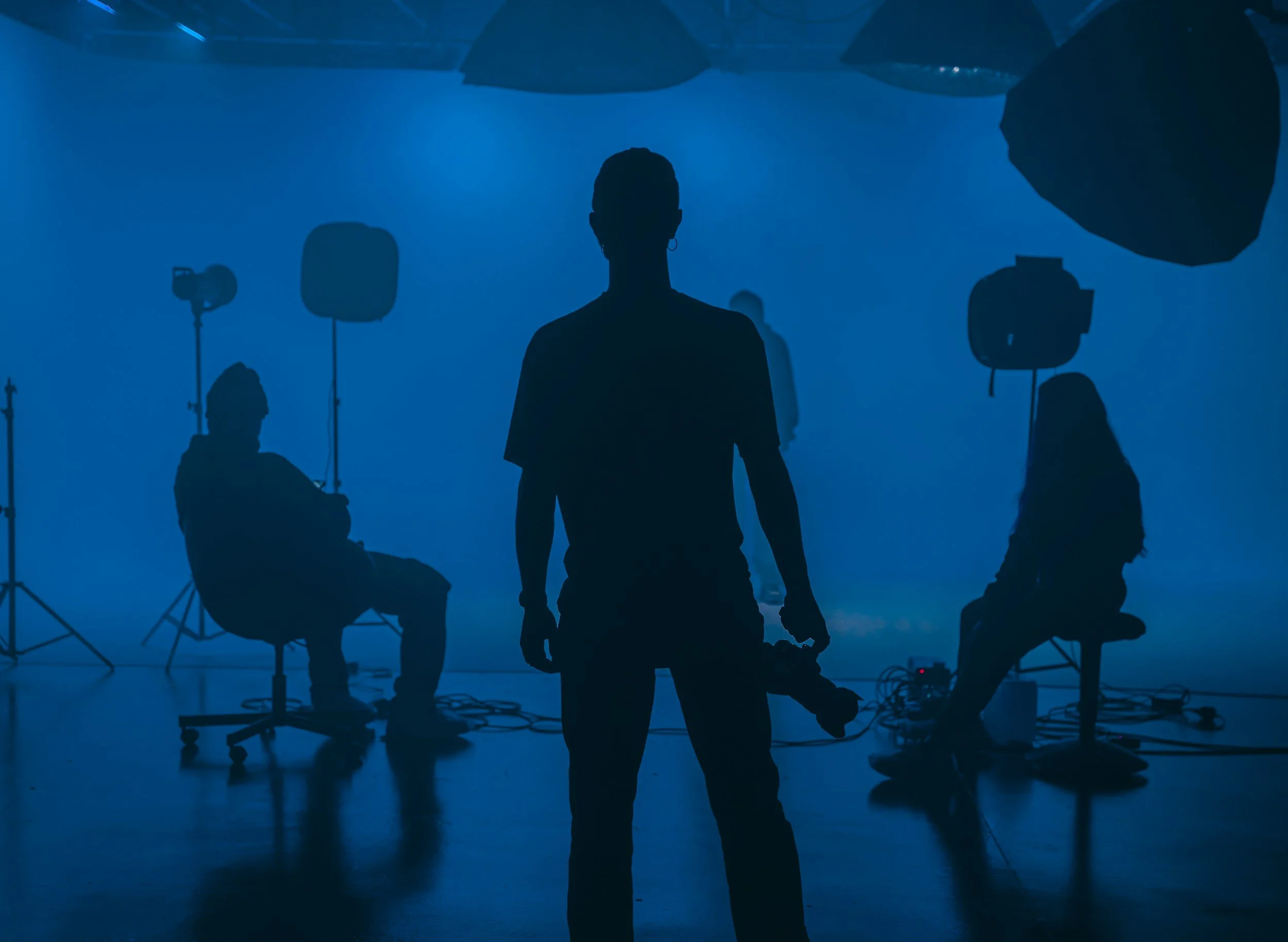 Silhouette of a person standing in a photography studio with professional lighting equipment and assistants working around, backlit with blue lighting.