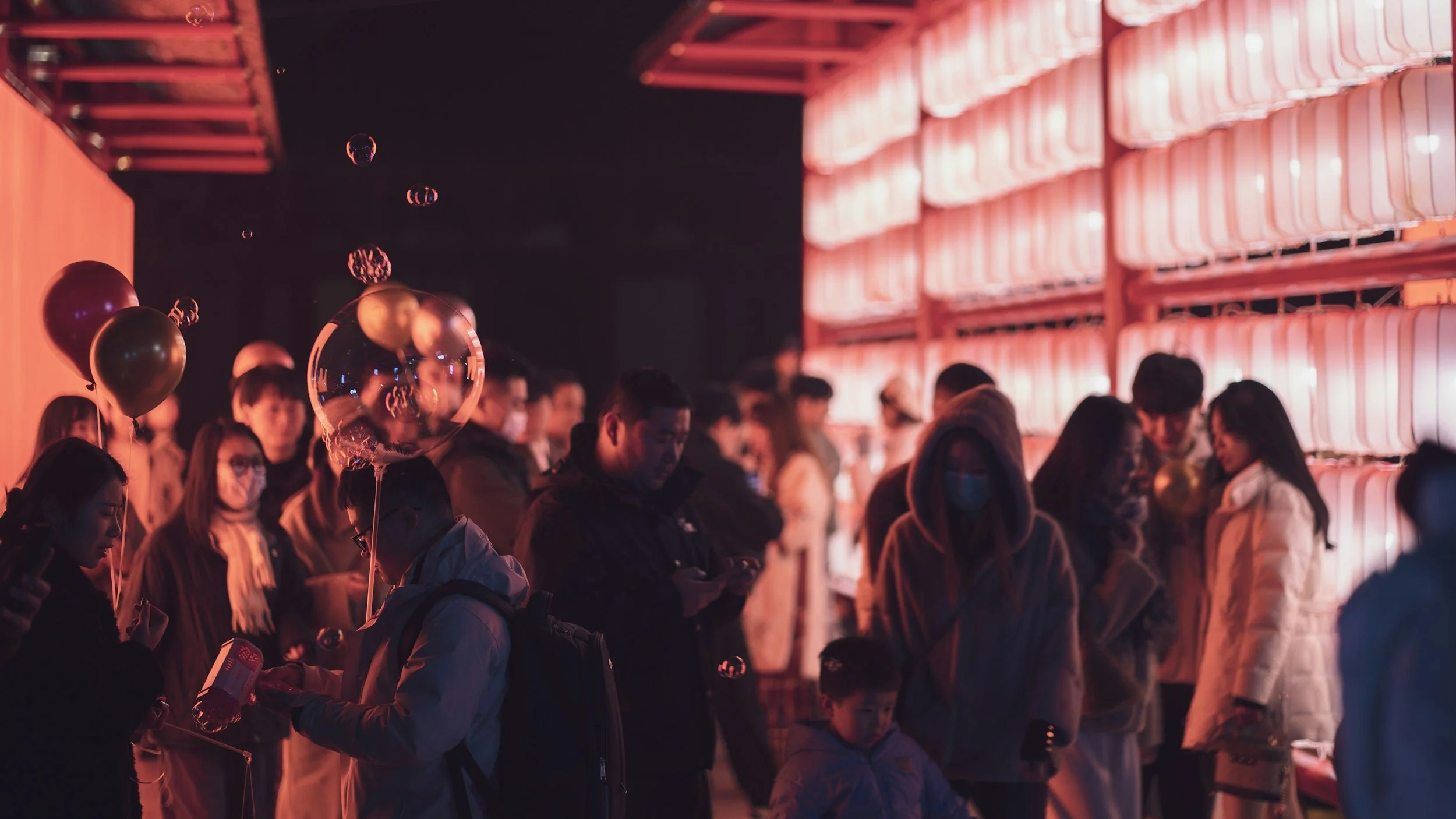 People standing in line at night next to a wall with illuminated pinkish-red panels, some holding balloons, some looking at their phones, in an outdoor event or gathering.