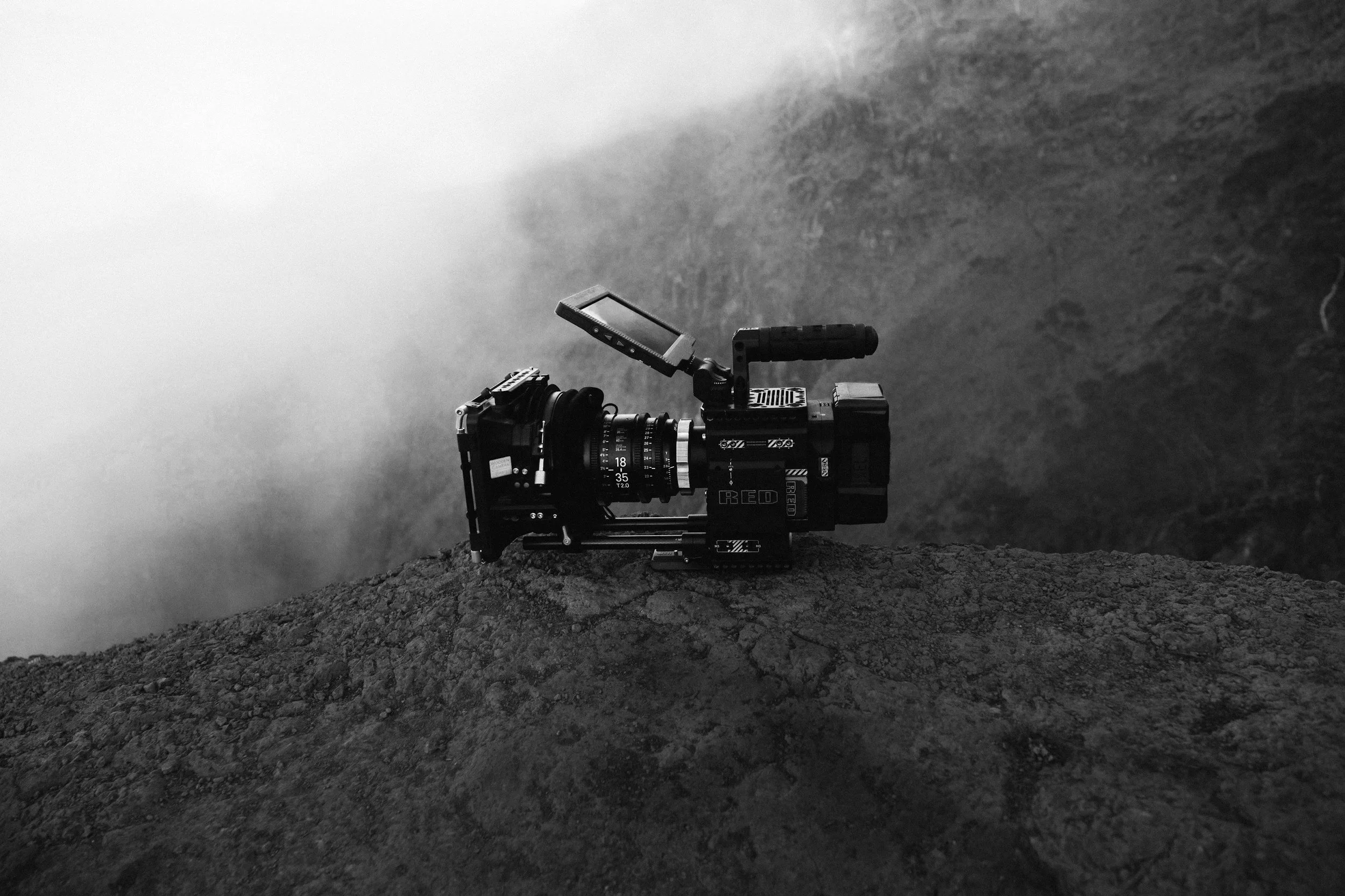 A professional camera mounted on a rugged surface, set against a misty or foggy landscape with fog obscuring the background.