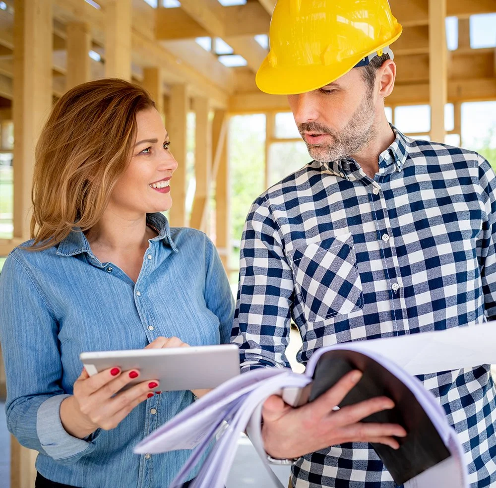 Two construction workers, a woman and a man wearing a yellow hard hat, are inspecting construction plans at a building site.