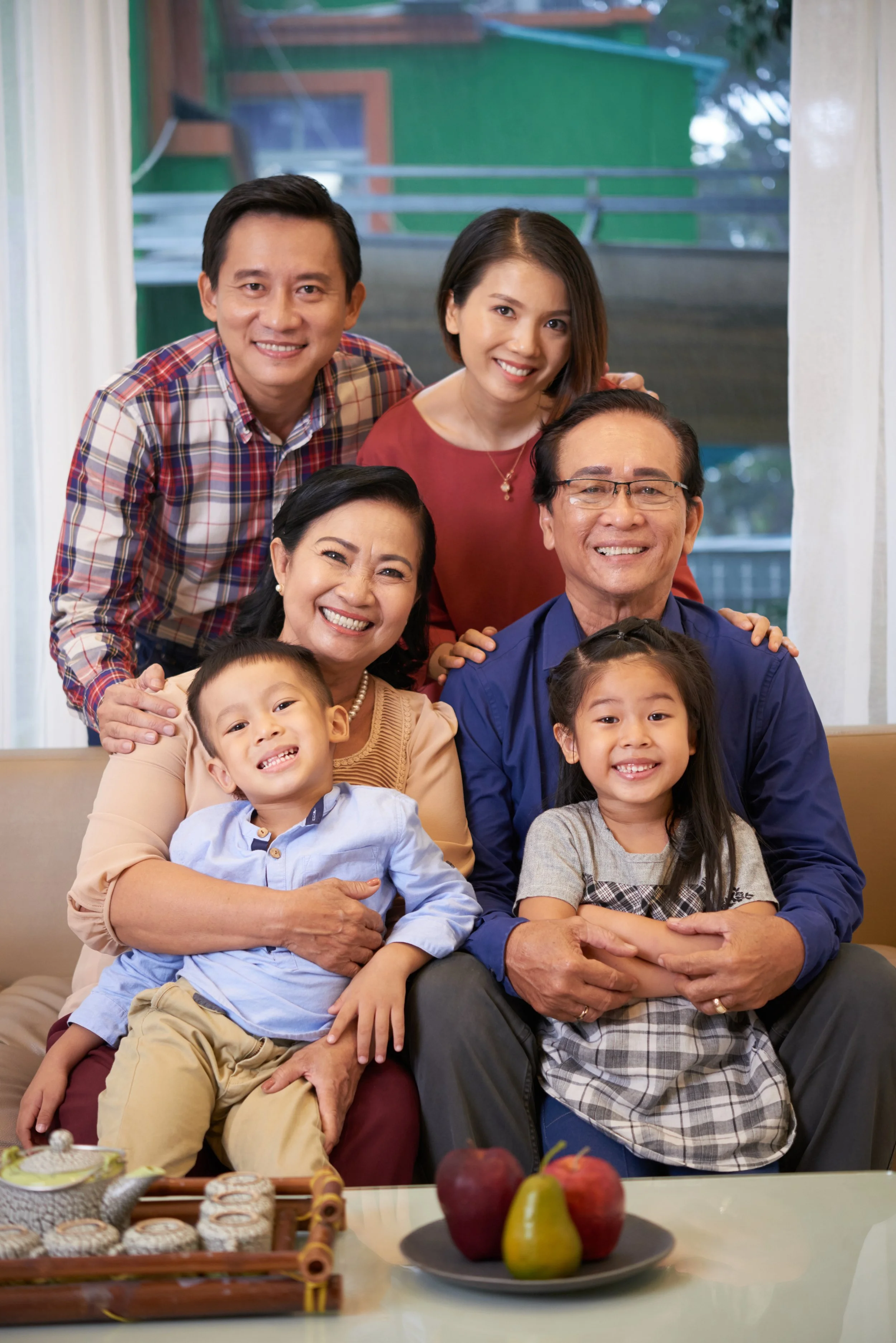 Family of seven gathered together indoors, smiling for a photo. In front, two children, a boy and a girl, sit with parents; three adults stand behind them. The scene shows a cozy, cheerful family moment.