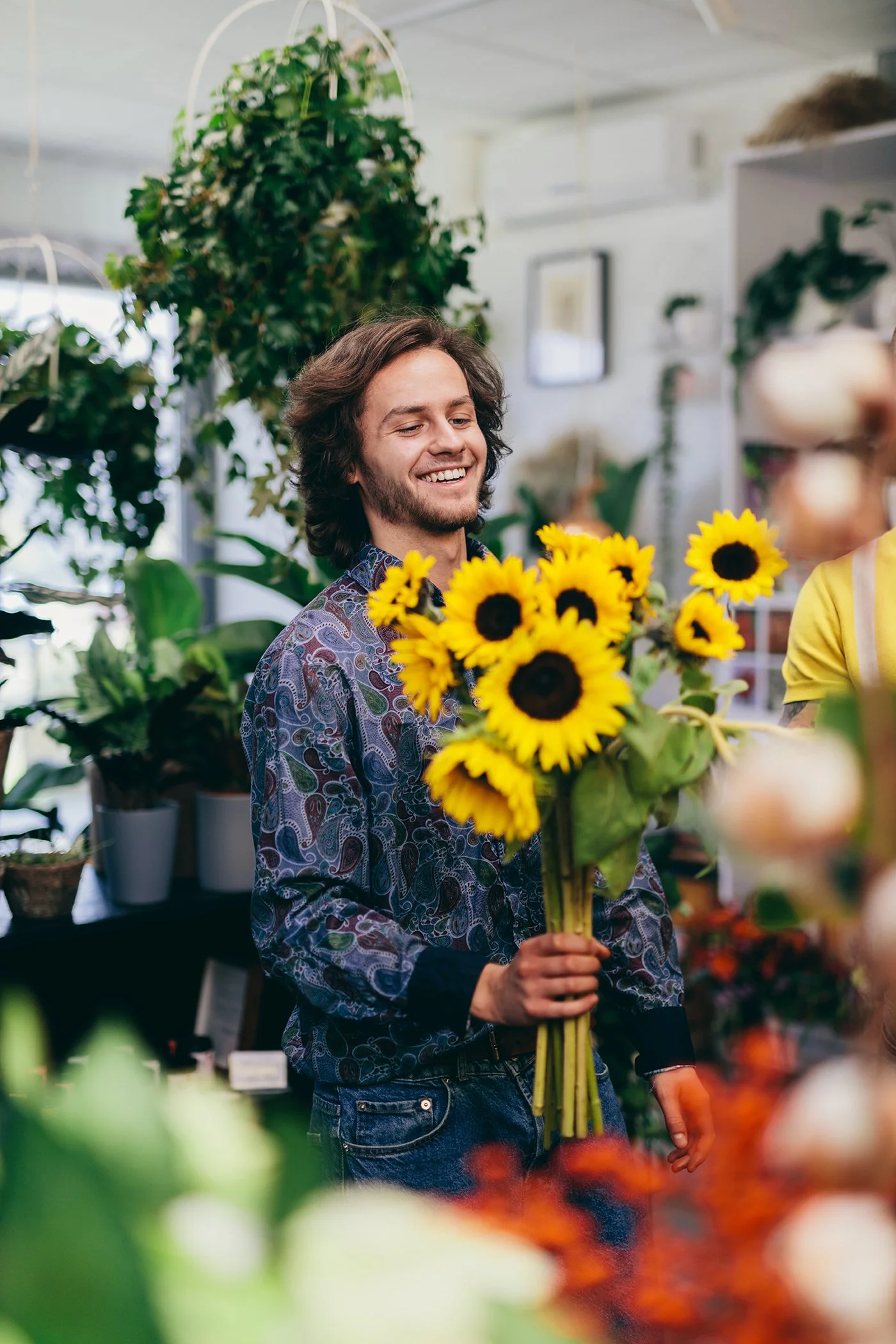 A young man with long brown hair and a beard is smiling and holding a large bouquet of bright yellow sunflowers inside a plant shop.
