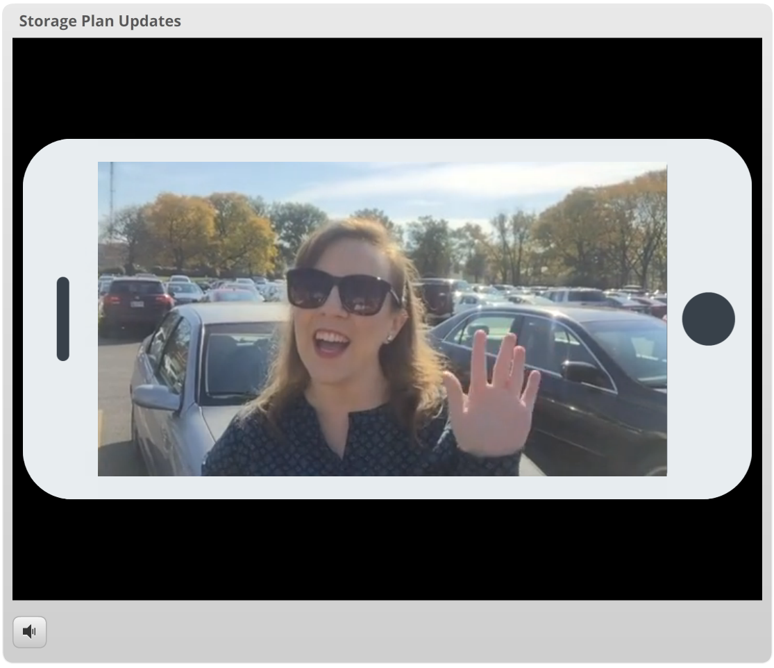 A woman wearing sunglasses and a patterned shirt, smiling and waving at the camera in a parking lot with cars and autumn trees in the background.
