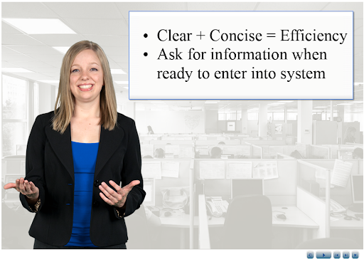 A woman standing in an office, smiling and gesturing with one hand, in front of a presentation slide that reads: 'Clear + Concise = Efficiency' and 'Ask for information when ready to enter into system'.