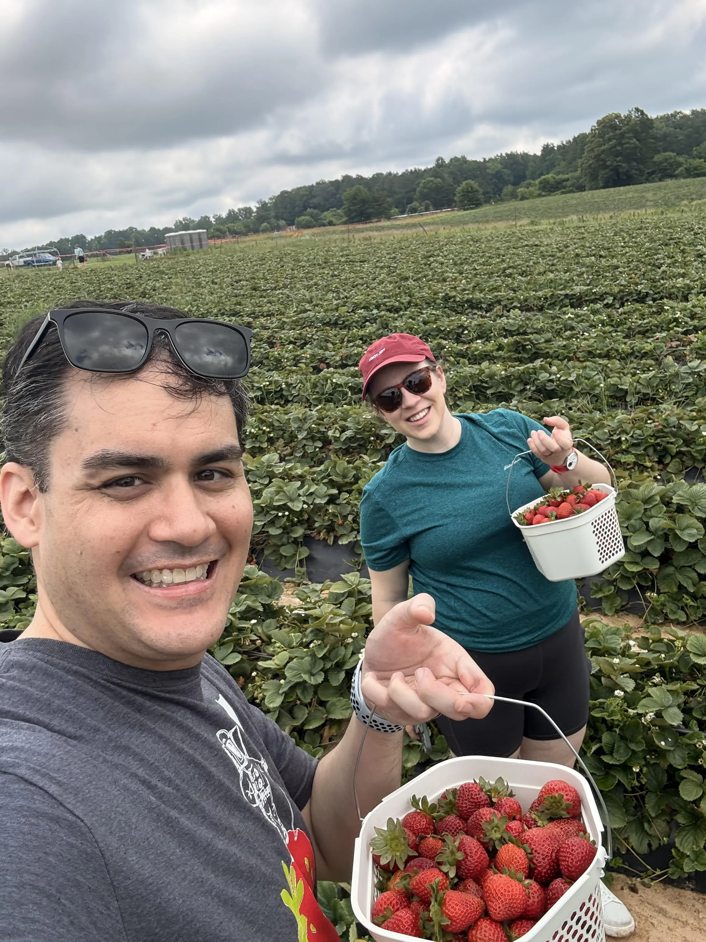 Two people smiling and holding baskets of strawberries in a strawberry field under cloudy sky.