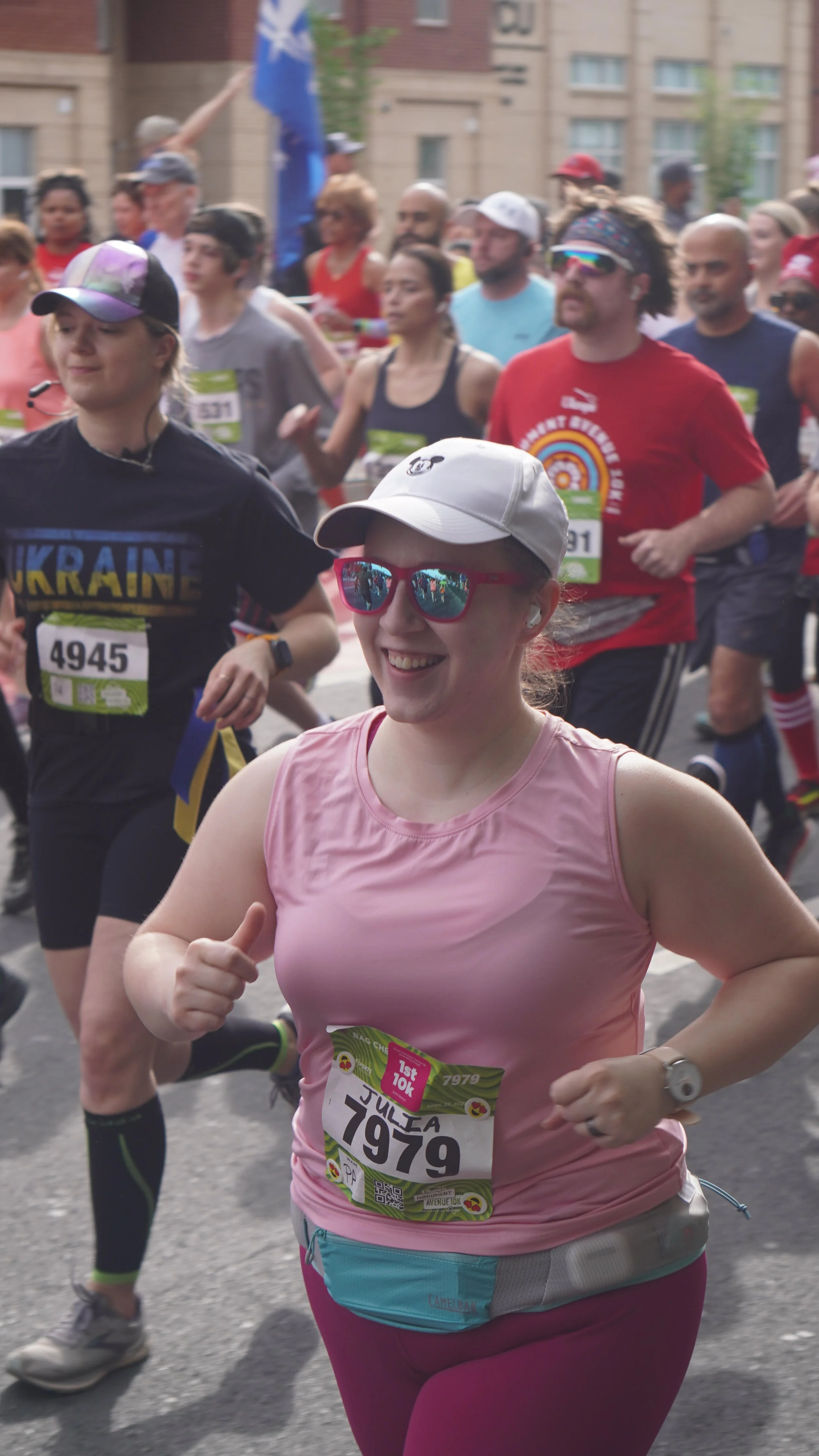 A group of runners participating in a marathon, with a woman in the foreground wearing a pink tank top, sunglasses, white cap, and a race bib number 1979, smiling and jogging in the street. Many other runners are visible behind her.