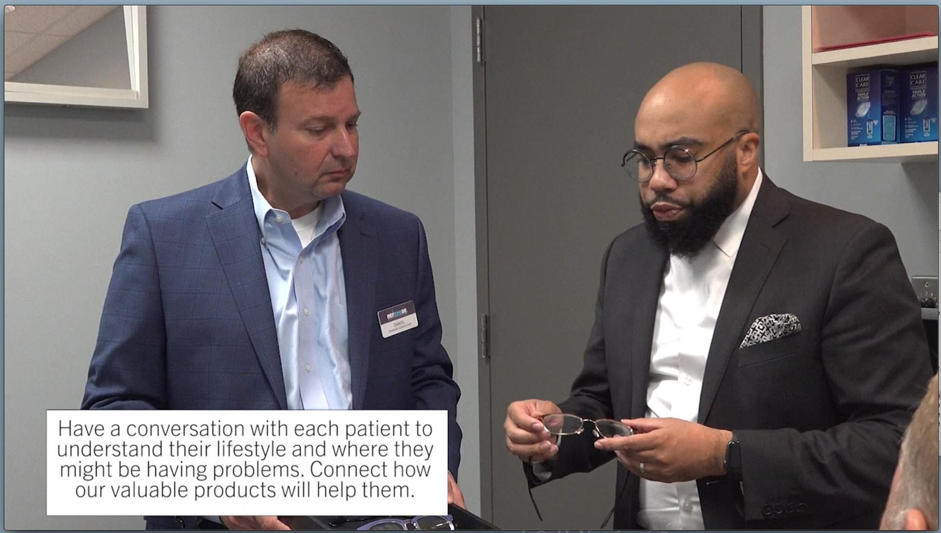 Two men in business attire are engaged in a conversation in an office setting. One man is holding a pair of glasses, and a caption reads, 'Have a conversation with each patient to understand their lifestyle and where they might be having problems. Connect how our valuable products will help them.'