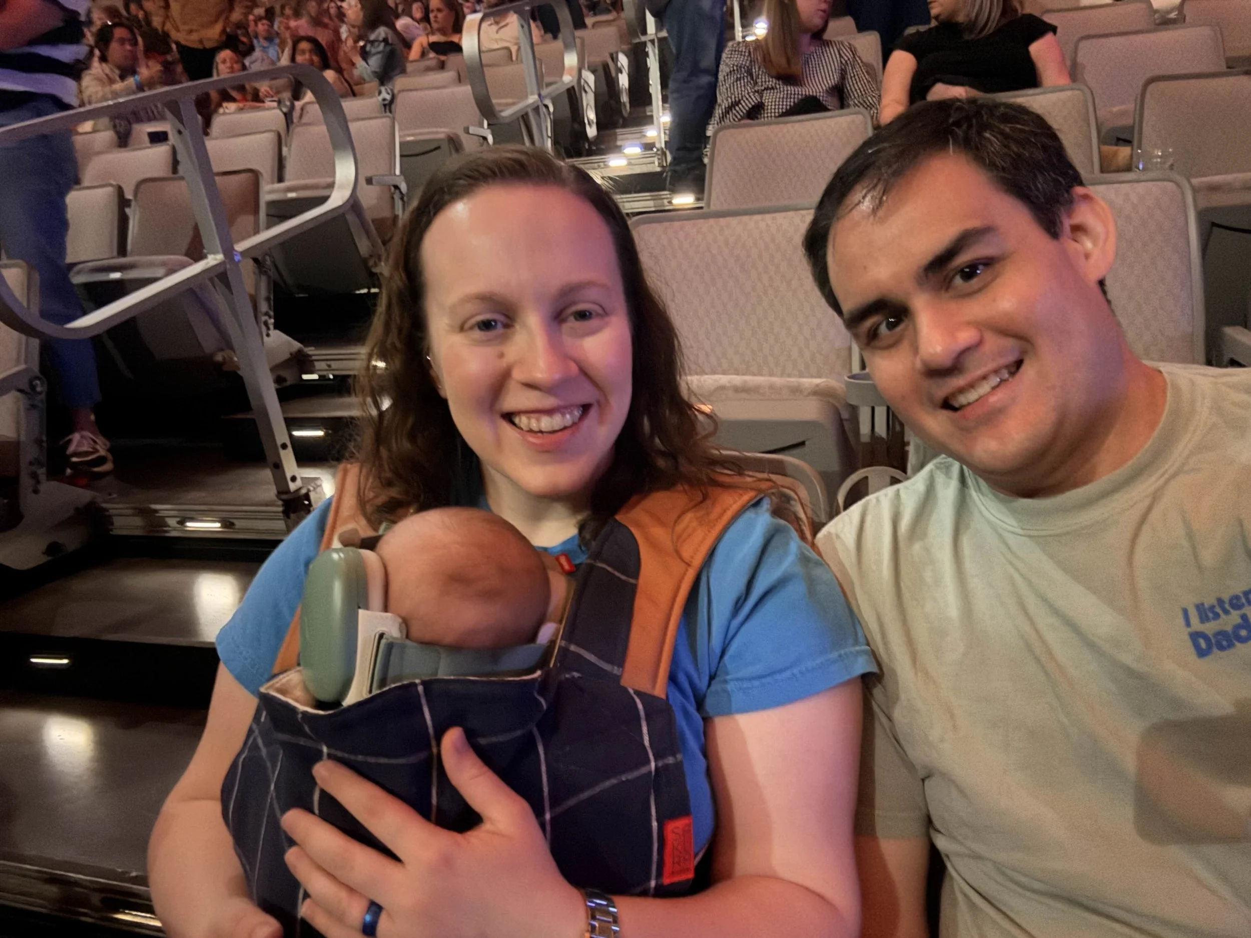 A happy family at an indoor arena, with a woman holding a baby in a carrier, smiling at the camera, and a man next to her smiling.